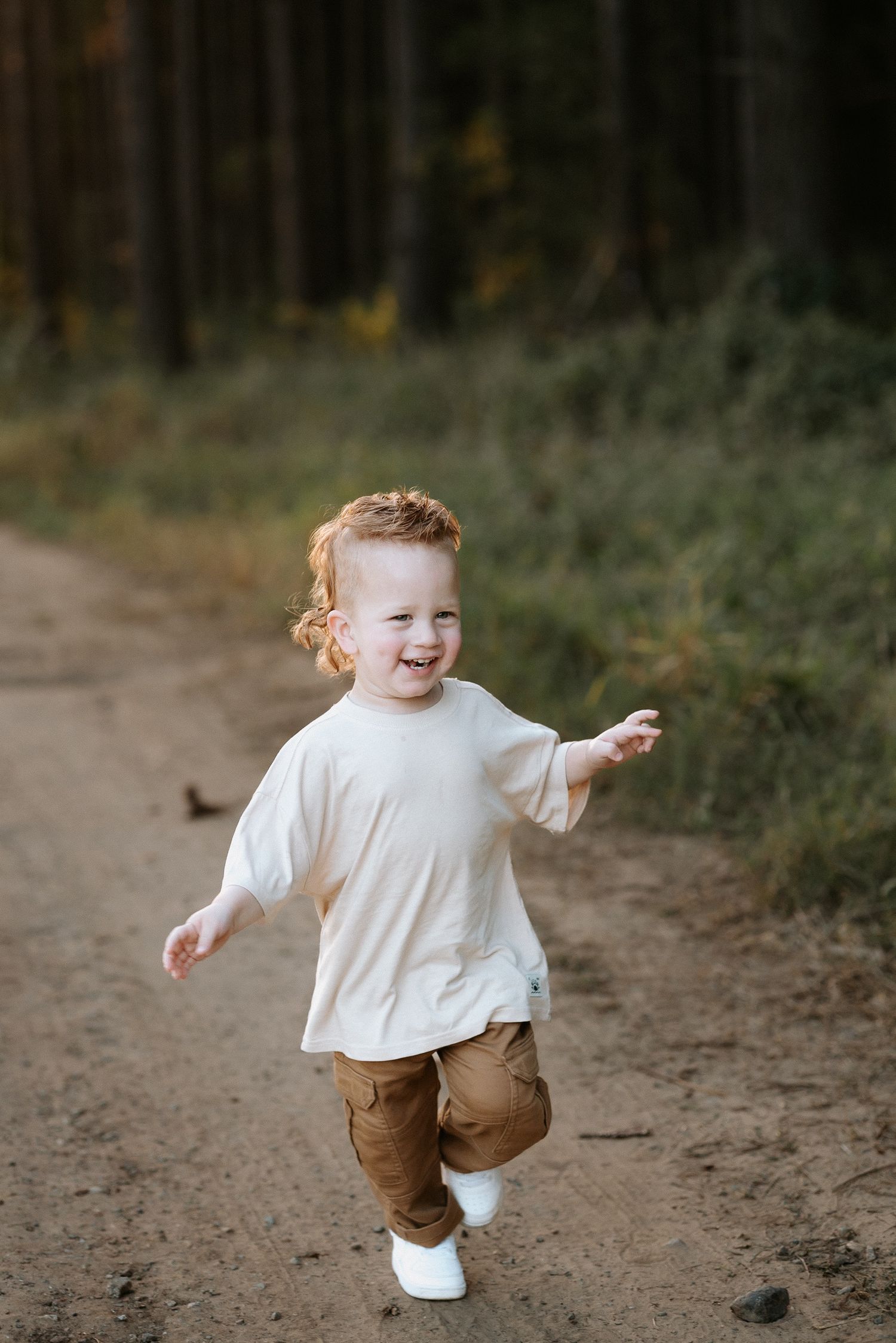 A joyful red-haired toddler in a white t-shirt and brown pants runs along a forest path, laughing with arms outstretched.