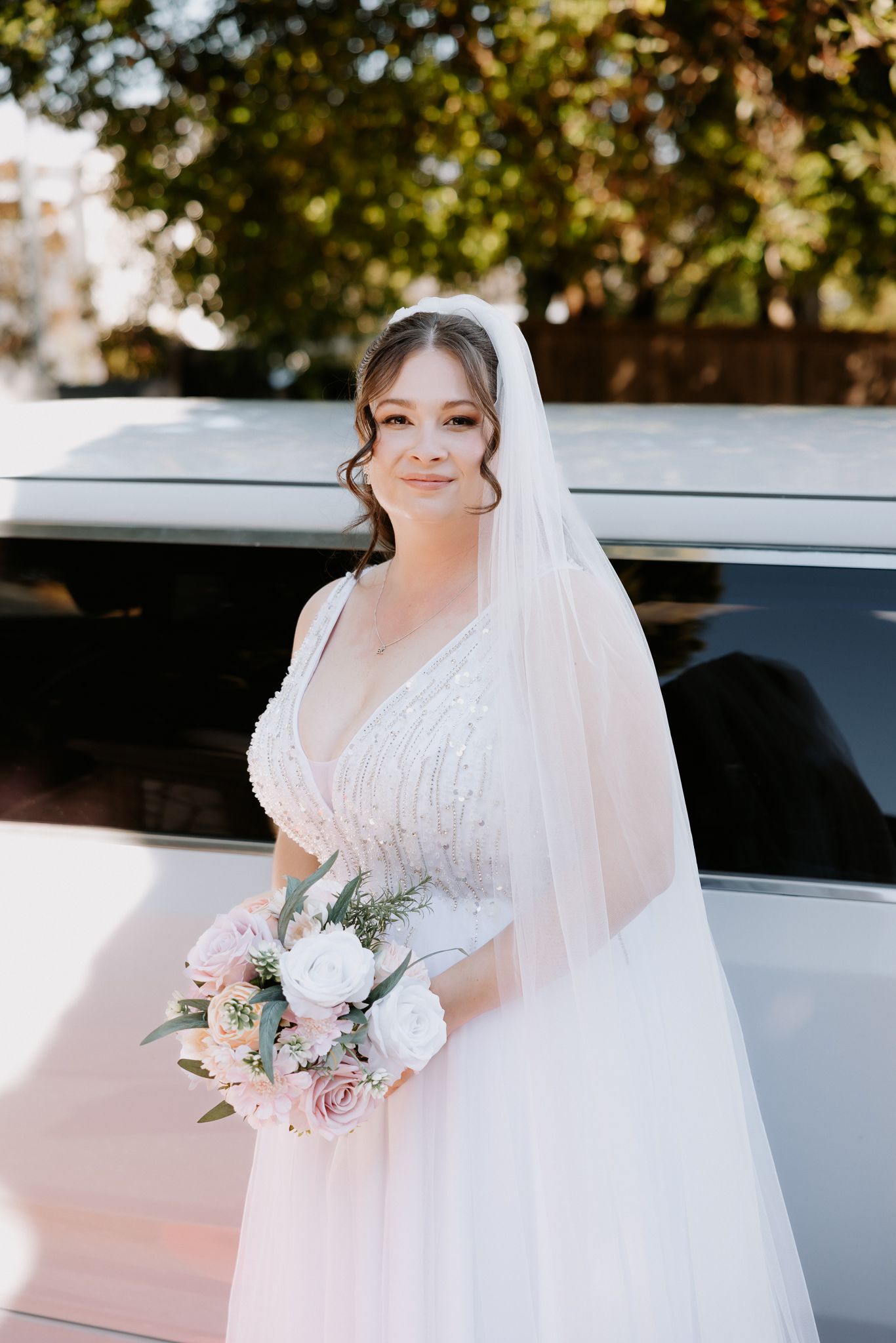 Bride in white beaded gown and veil holds pink and white bouquet, standing beside a white limousine outdoors.