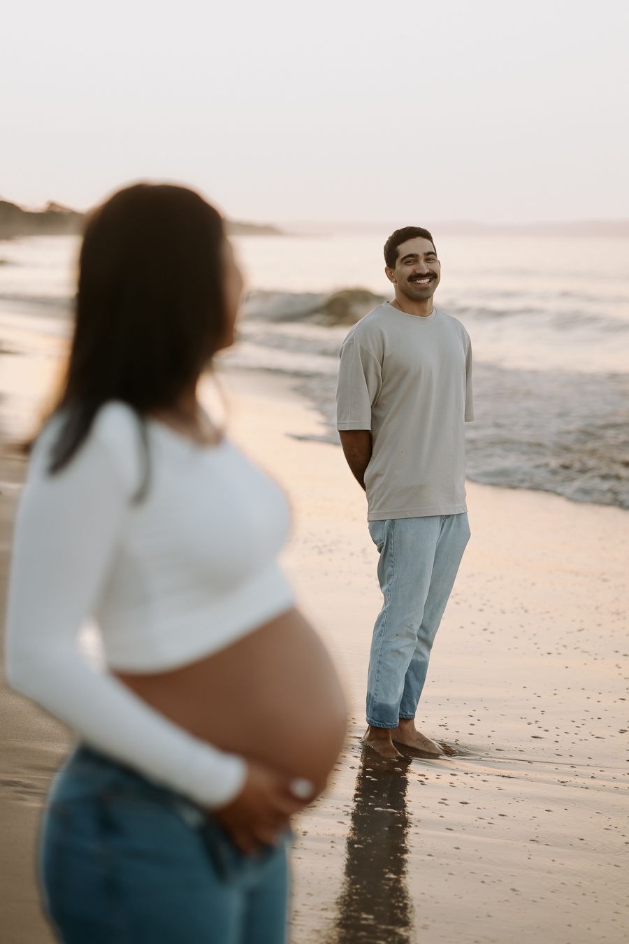 A couple stands on a beach at sunset, with one person showing their pregnant belly in a white sweater and jeans.