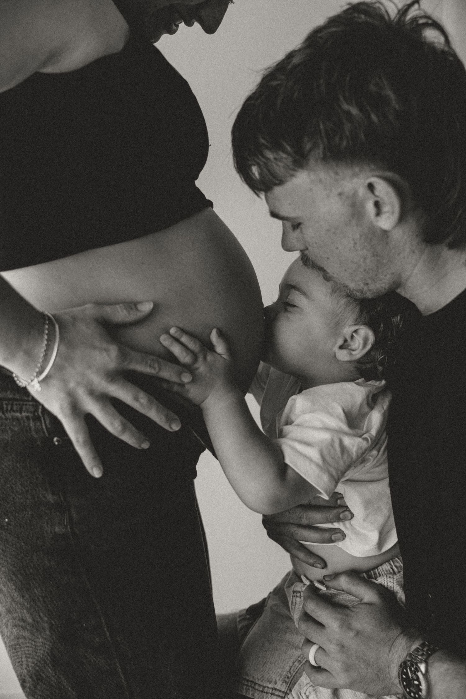 A black and white photograph shows a young child touching a pregnant belly while being held in an intimate family moment.