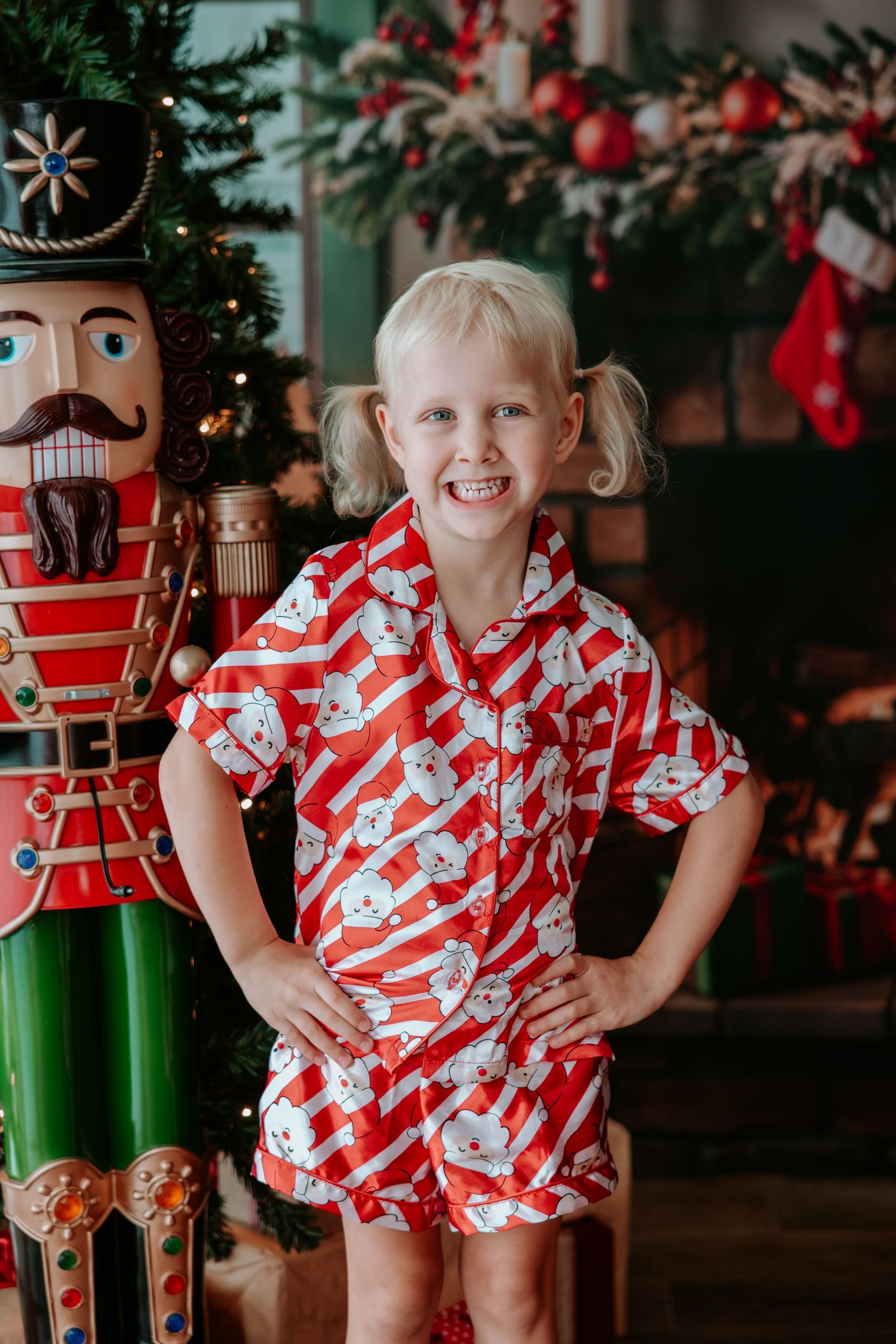 Young girl in red Christmas pajamas smiling beside a nutcracker decoration and holiday tree.
