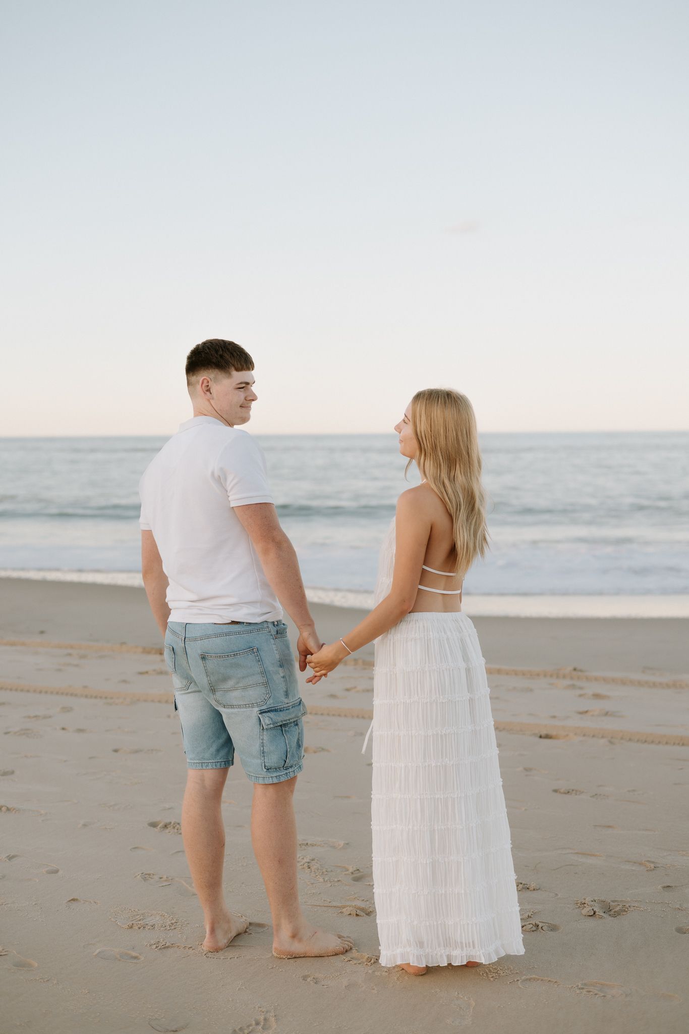 Couple holding hands on a sandy beach at sunset, woman in white maxi dress and man in blue shorts.