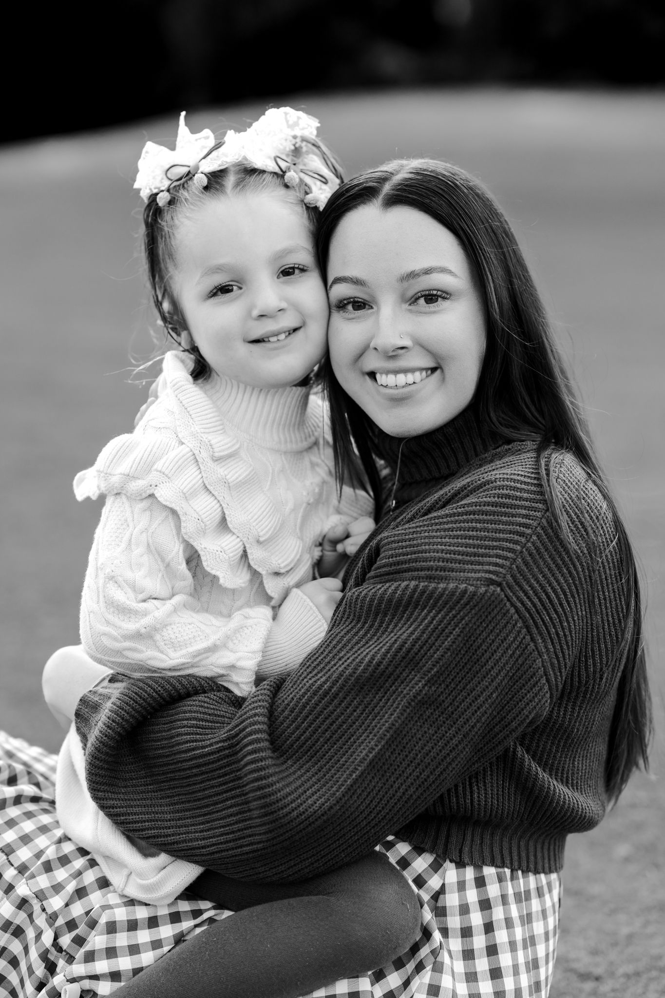 Black and white photo of a smiling woman holding a young girl with flower hair accessories outdoors.