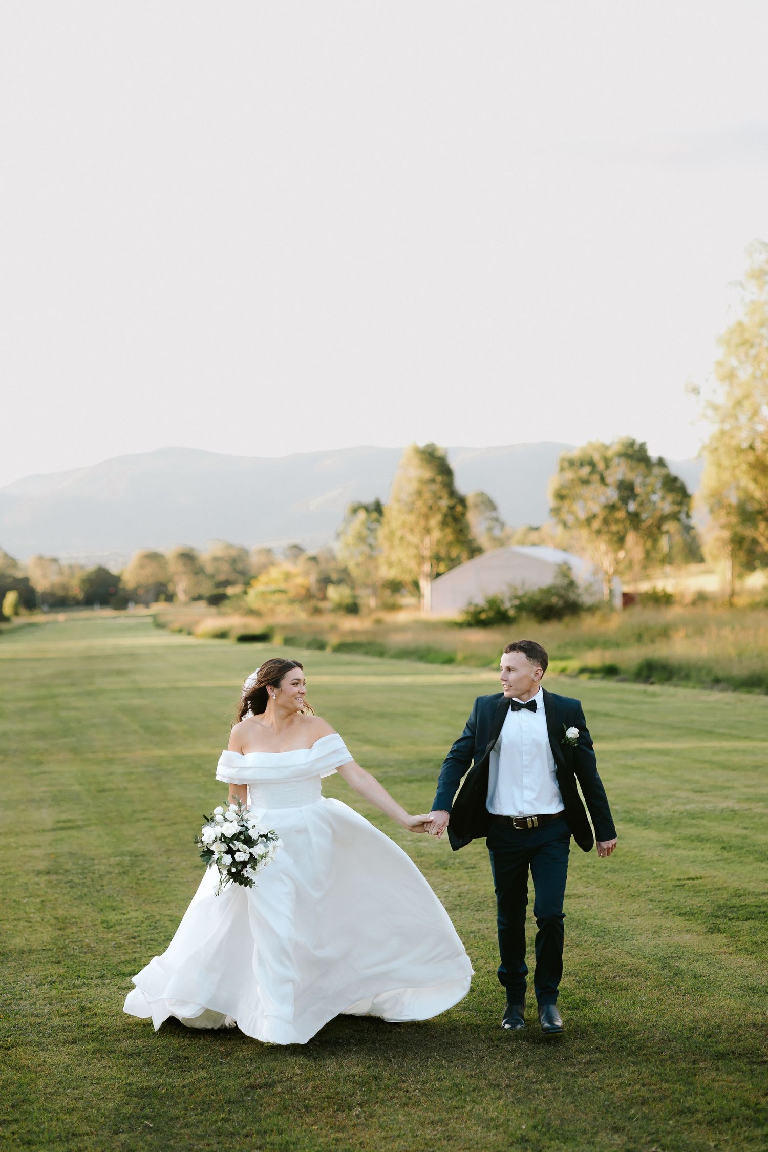 Bride and groom hold hands walking across green lawn with mountain backdrop on their wedding day.