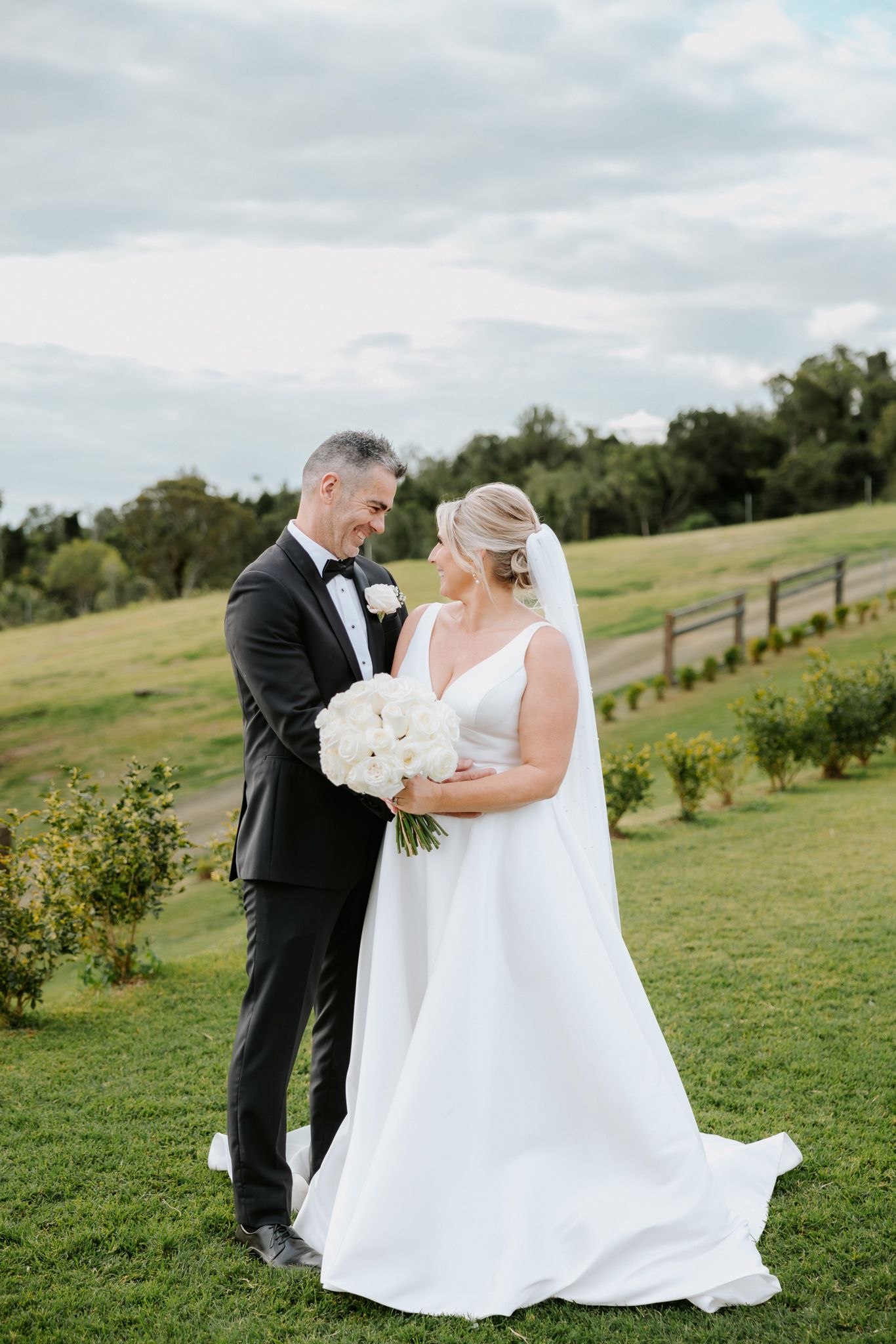 Bride and groom share a romantic moment on lush green lawn, bride holding white bouquet in elegant gown.