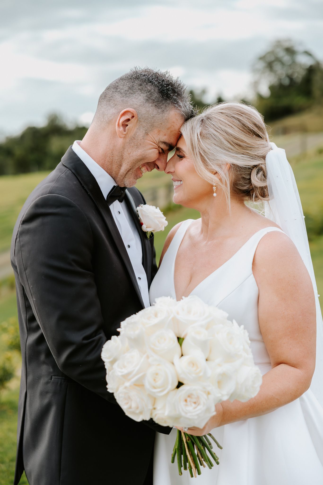 Bride and groom touching foreheads and smiling, bride holding white rose bouquet, outdoors on wedding day.
