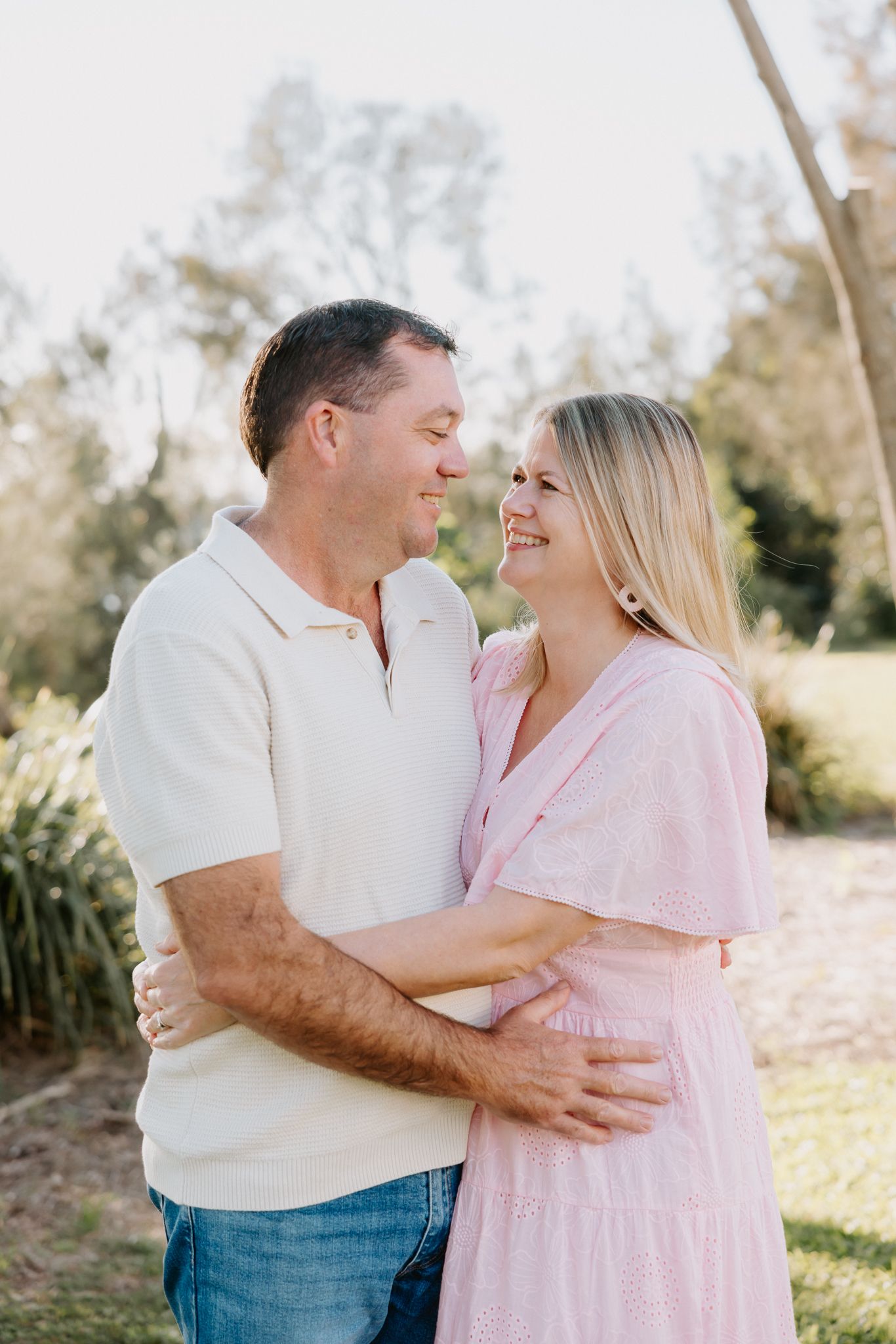 A smiling couple embraces outdoors, woman in pink dress and man in cream polo, sharing a romantic moment in a sunny park.