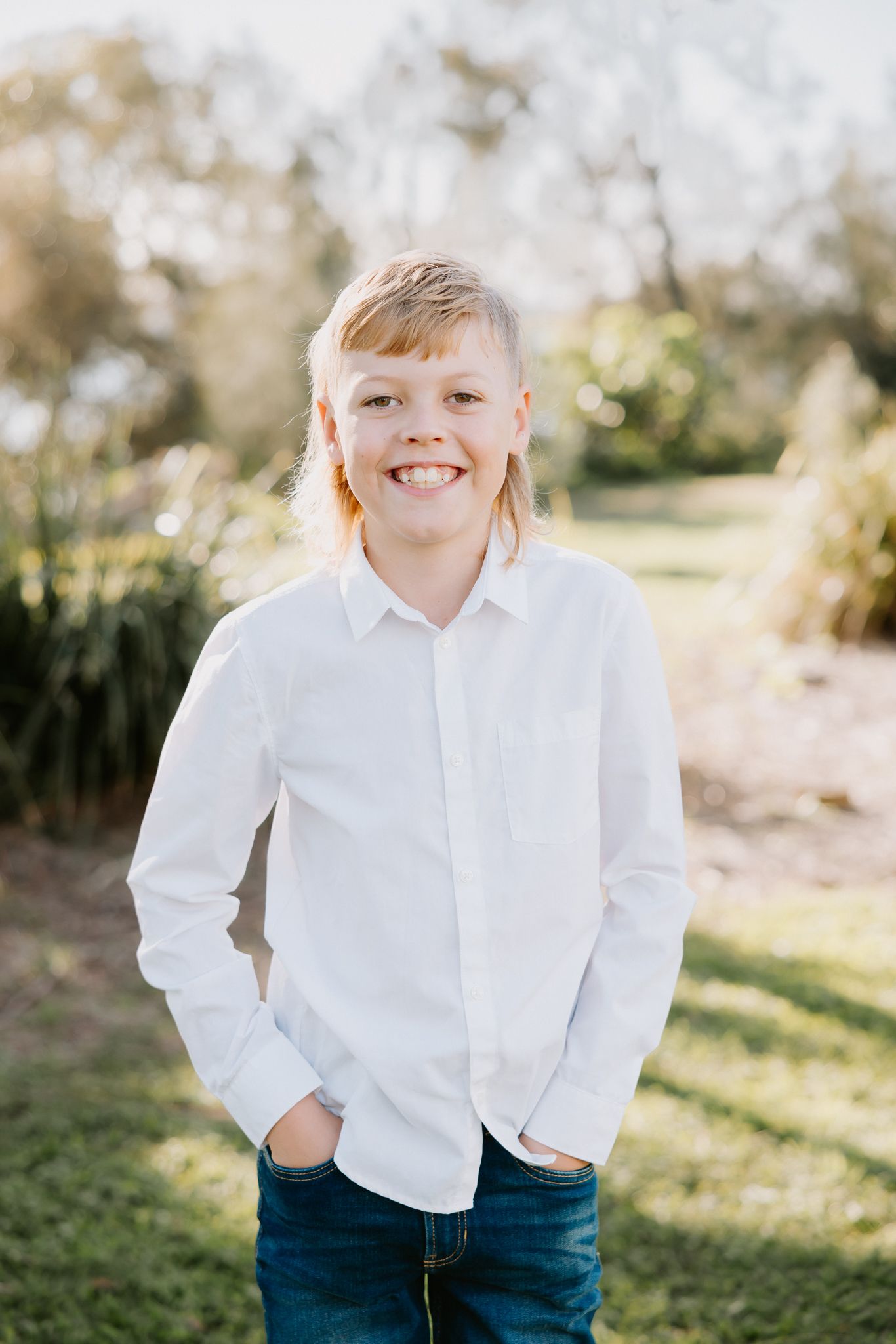 Smiling young boy in white button-up shirt and jeans standing outdoors in a sunny green park setting.