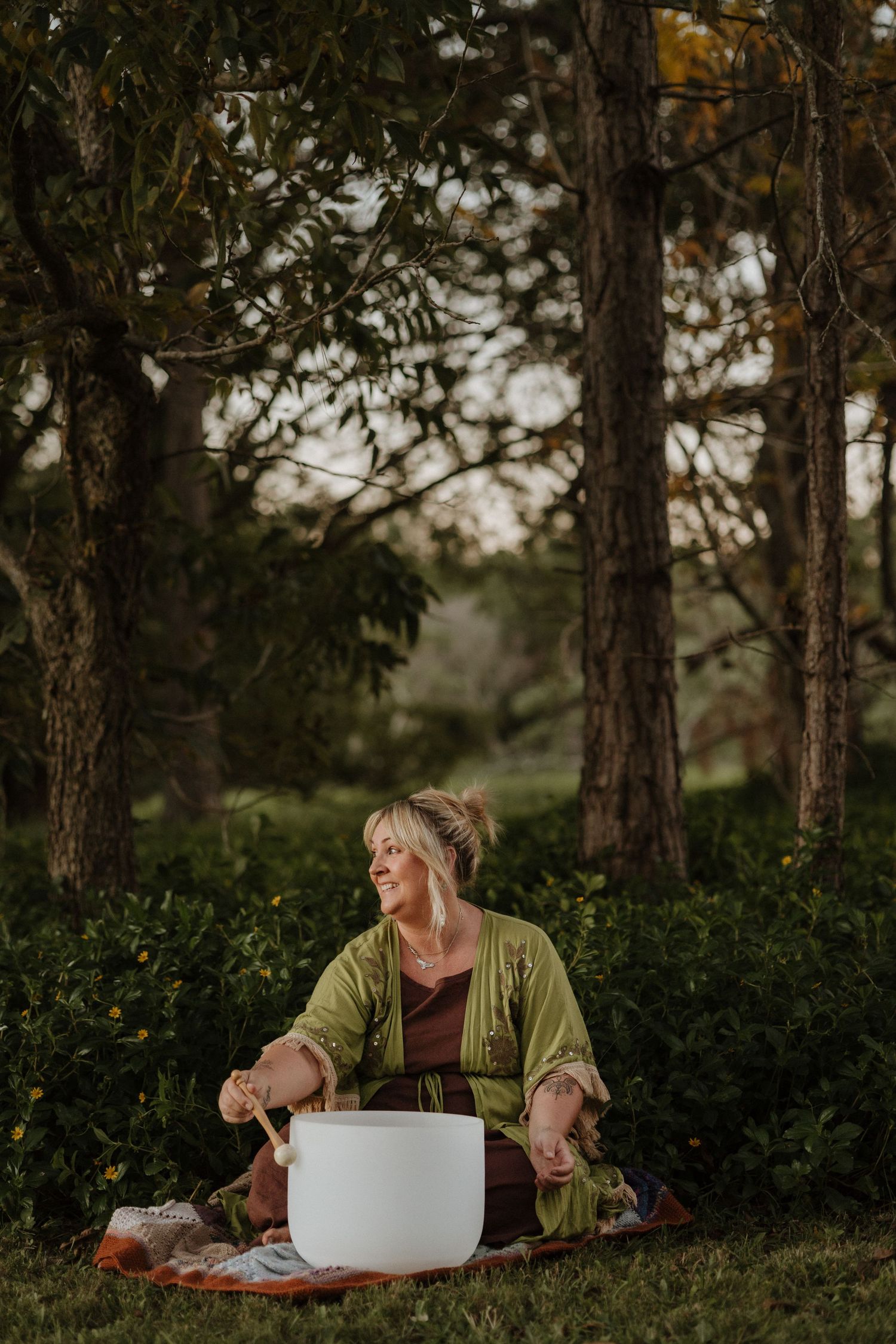 Woman playing a white crystal singing bowl outdoors, seated on grass among trees in a lush green park setting.