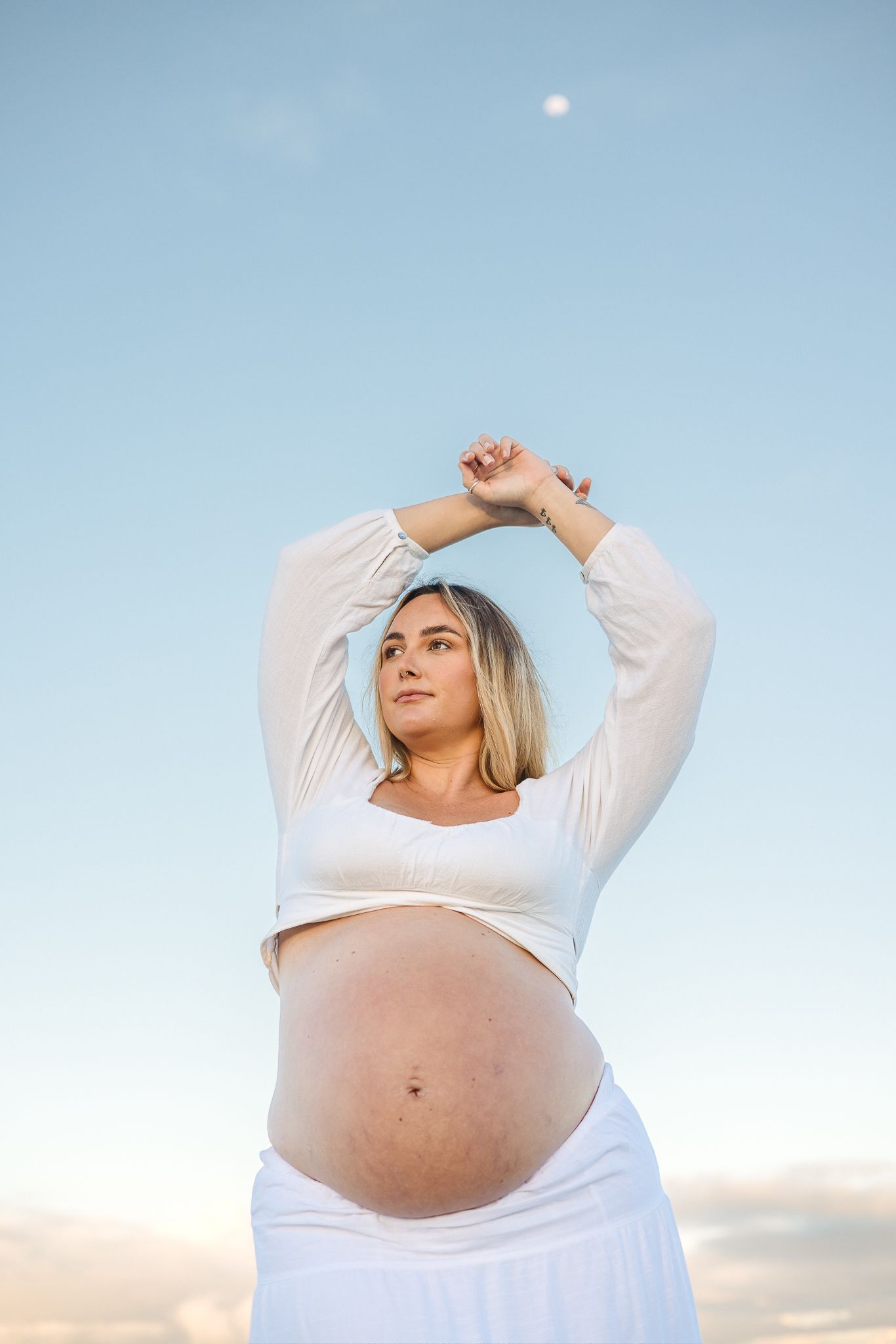 Pregnant woman in white crop top and skirt poses outdoors against blue sky, arms raised gracefully above her head.