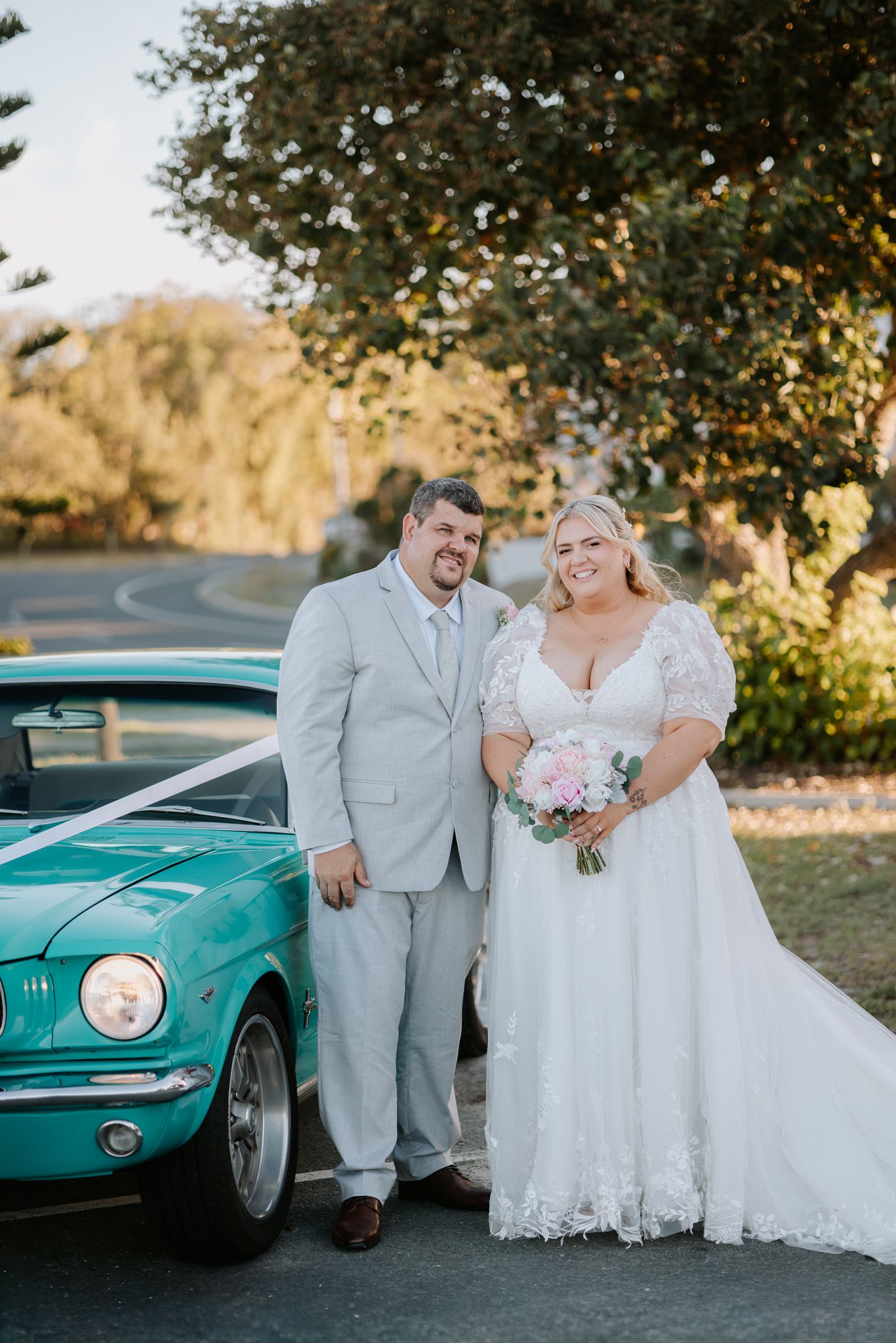 Bride and groom posing in wedding attire beside a vintage teal Ford Mustang decorated with white ribbon.