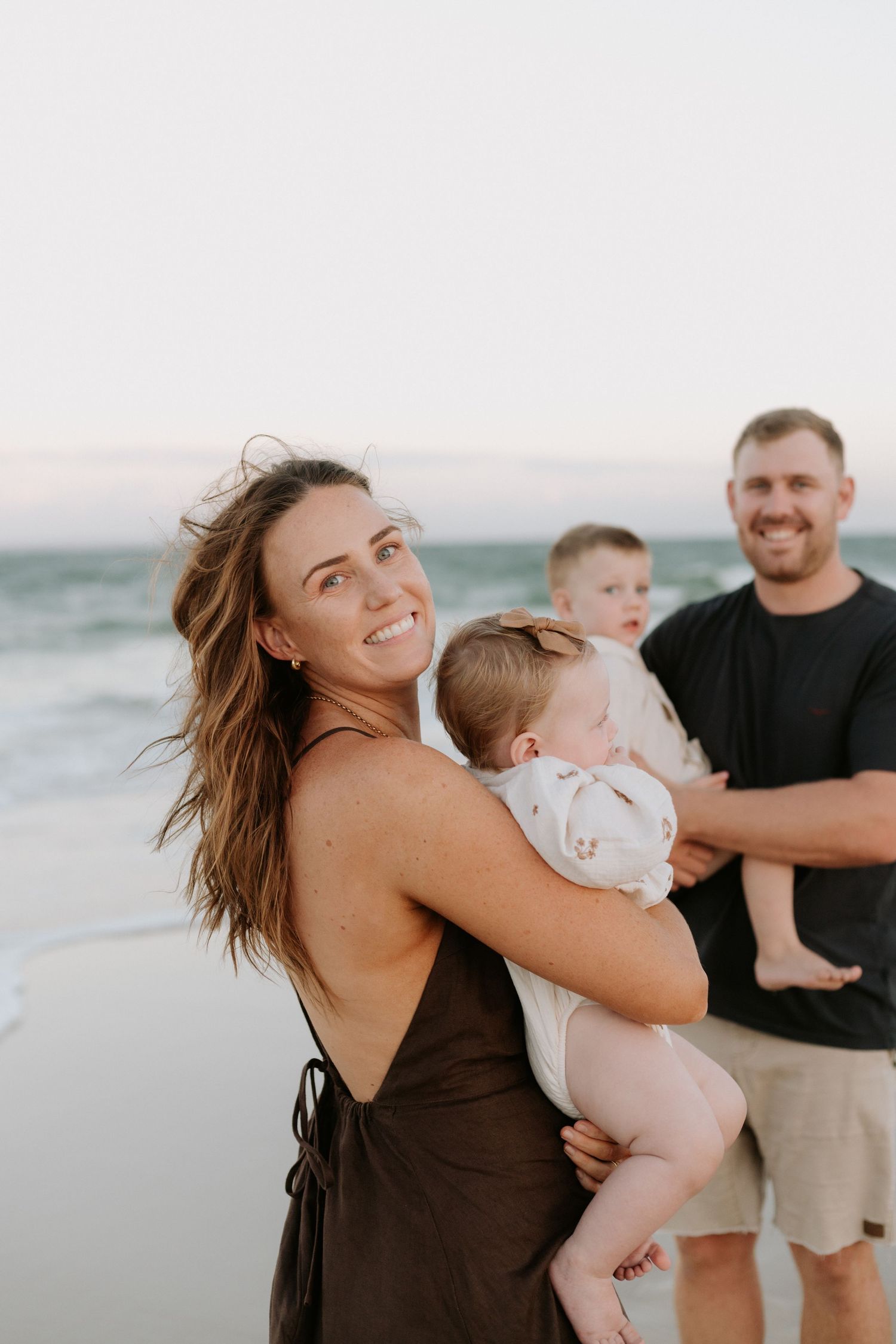 A smiling woman in a brown dress holds a baby on a beach, with a man and toddler in the background.