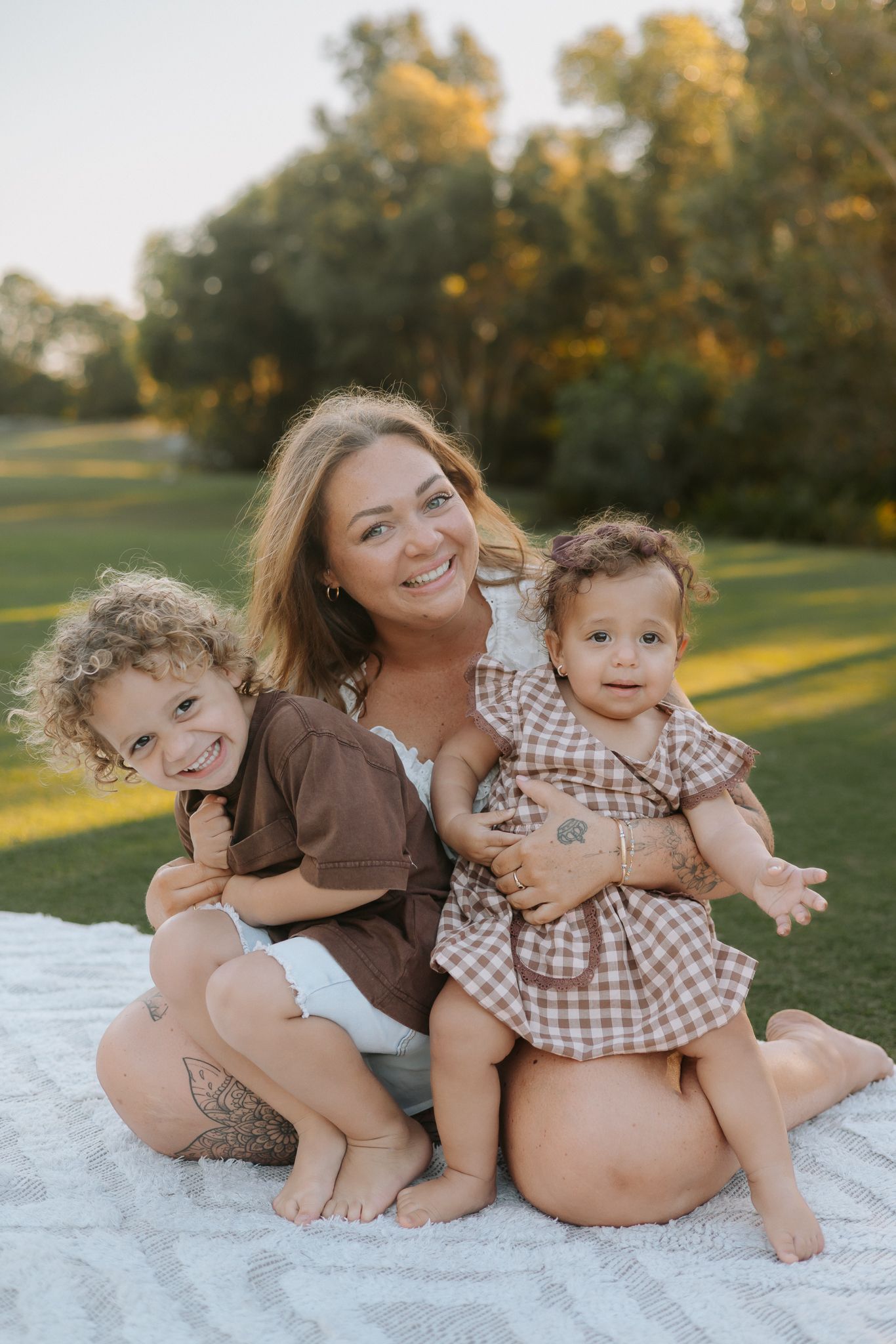A smiling woman sits on a blanket outdoors with two young children, surrounded by lush green trees in warm golden light.