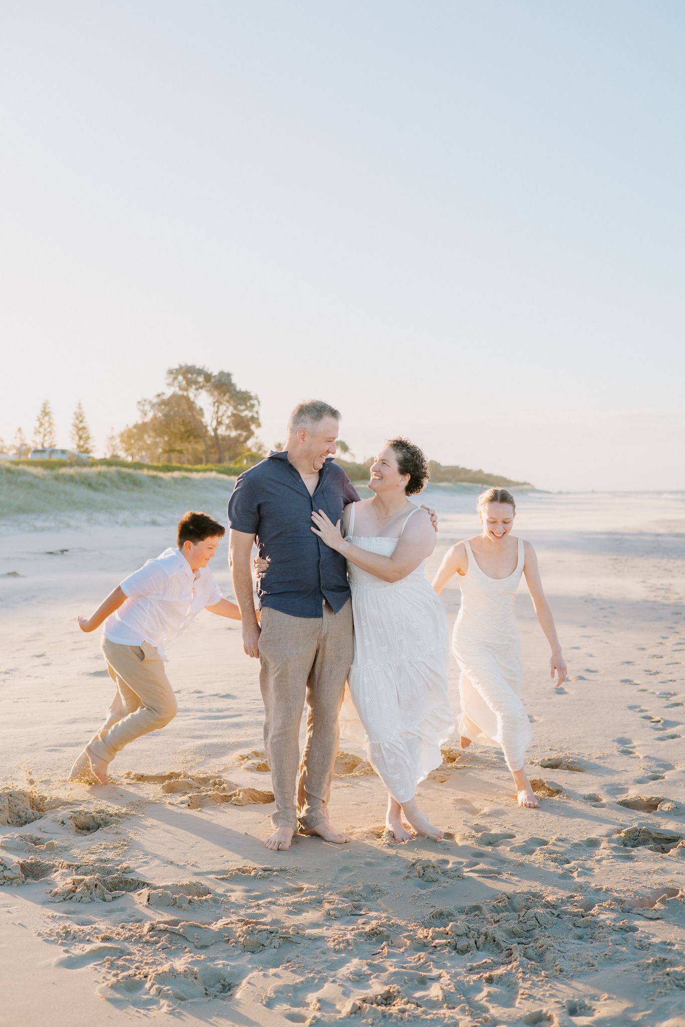 Family of four laughing and walking together on a sunny beach, parents embracing while children play around them.
