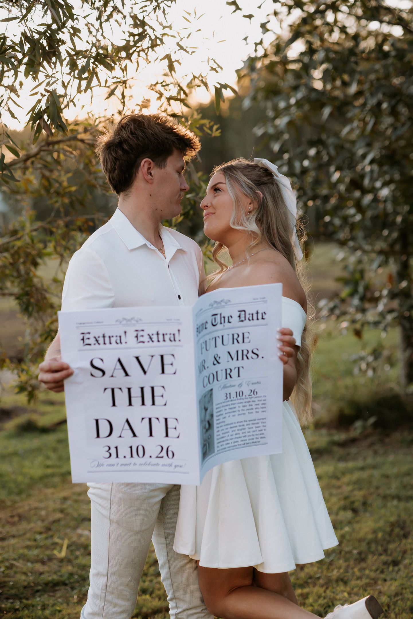 Couple holding newspaper-style Save the Date cards for 31.10.26 wedding, posing outdoors under olive trees.