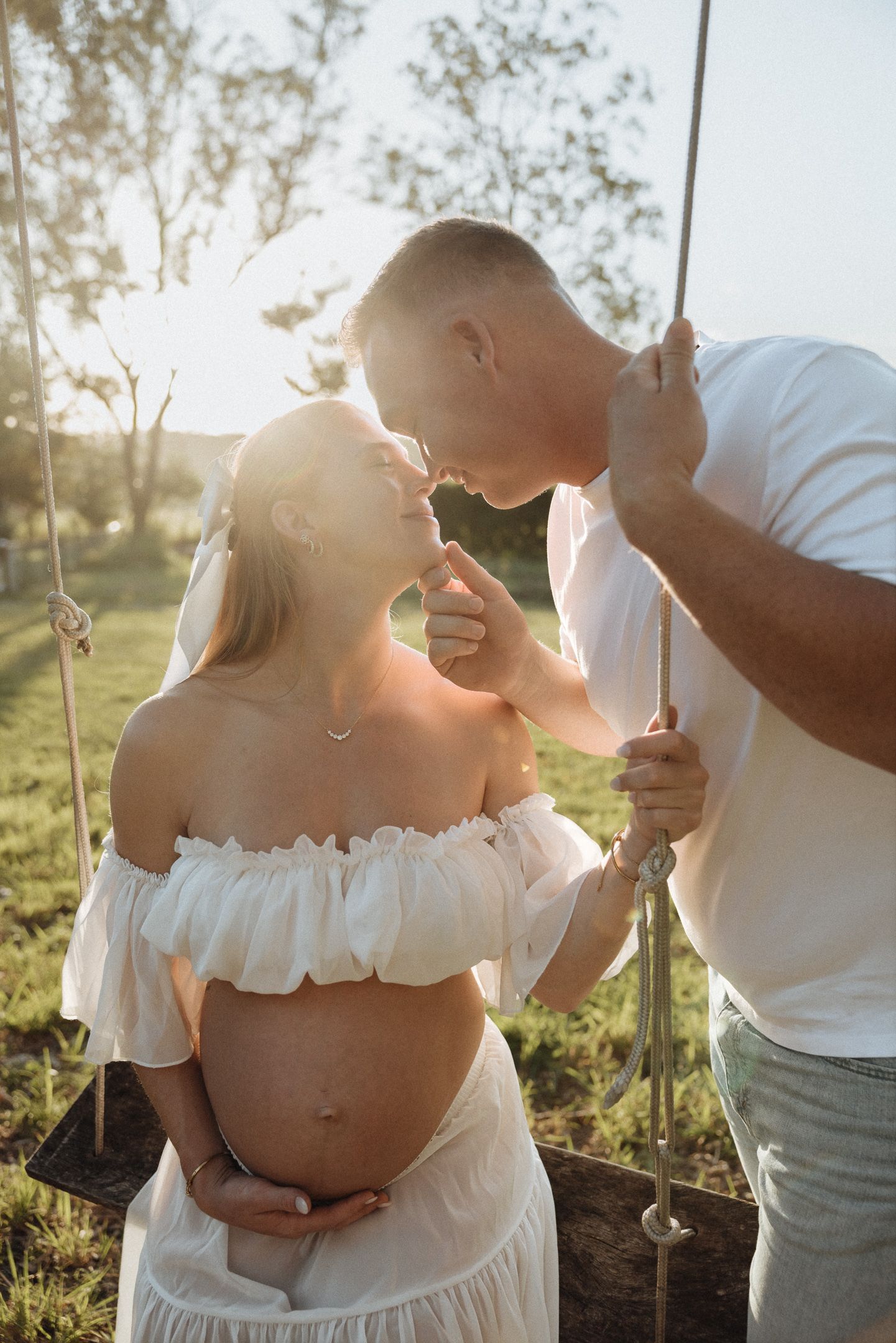 Pregnant couple sharing a tender moment on a swing at golden hour, woman in white off-shoulder dress cradling her baby bump.