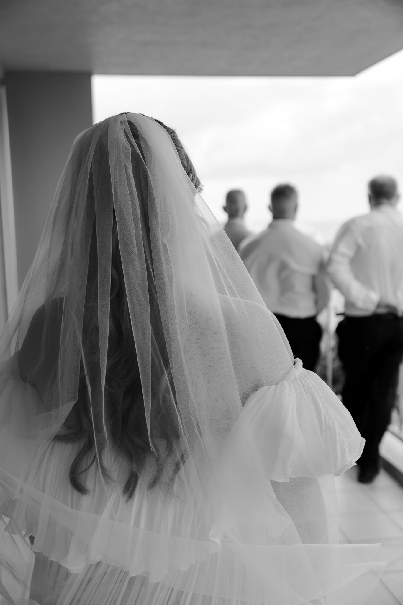 Black and white photo of a bride in veil and dress viewed from behind with groomsmen standing ahead.