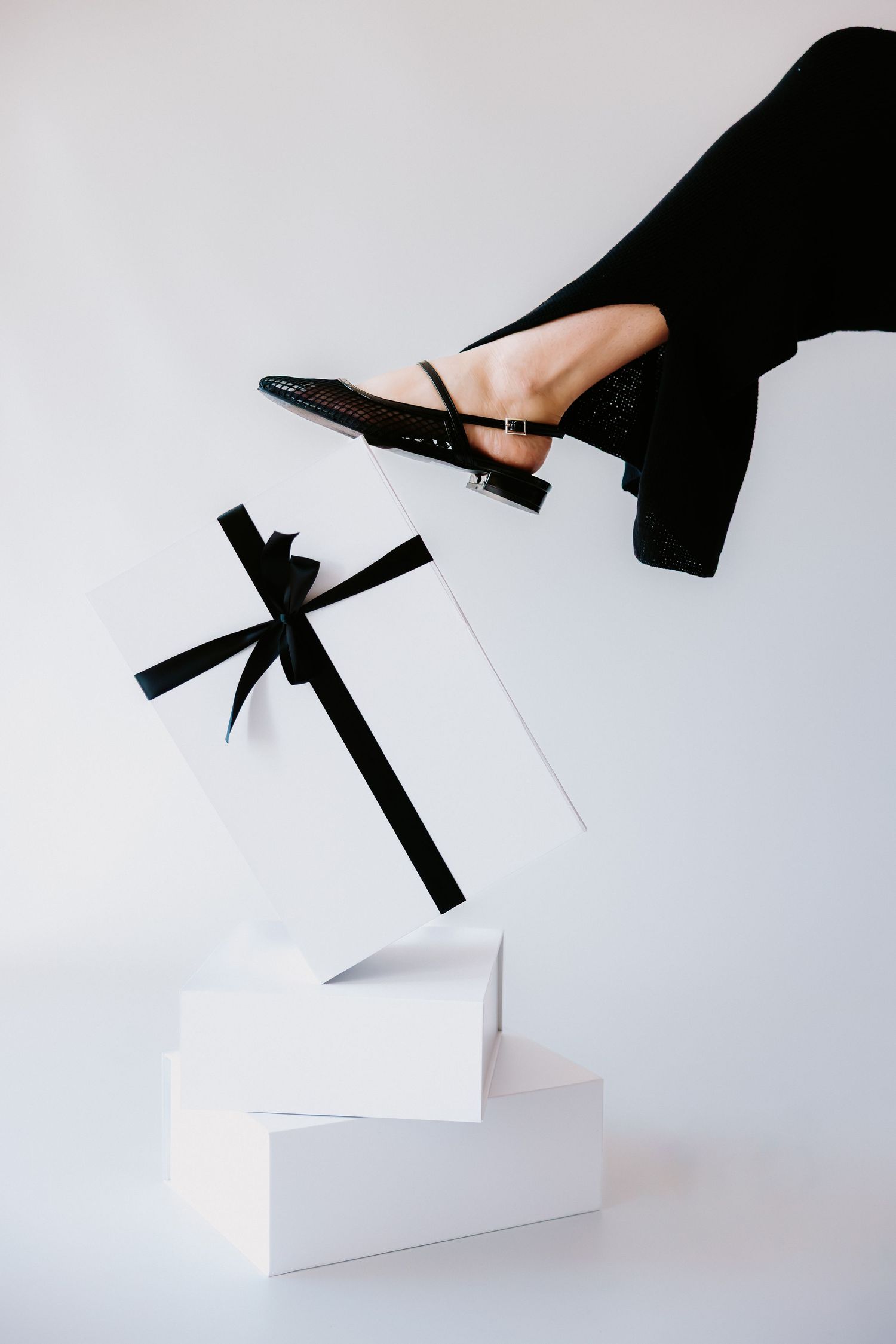 Elegant black heeled mule shoe on a leg in black dress beside white gift boxes with black ribbon bow.