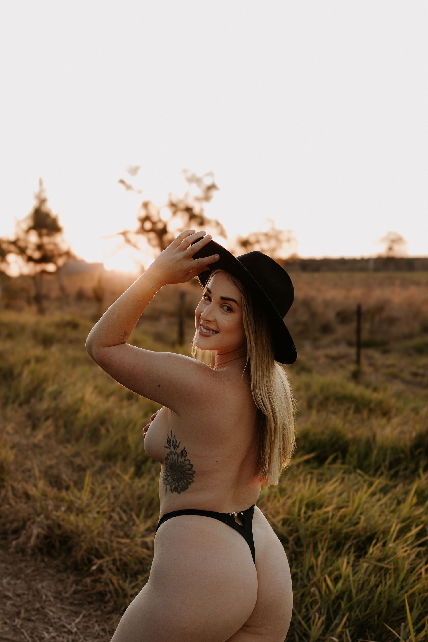 Blonde woman in black hat and thong posing outdoors at golden sunset in a grassy field.