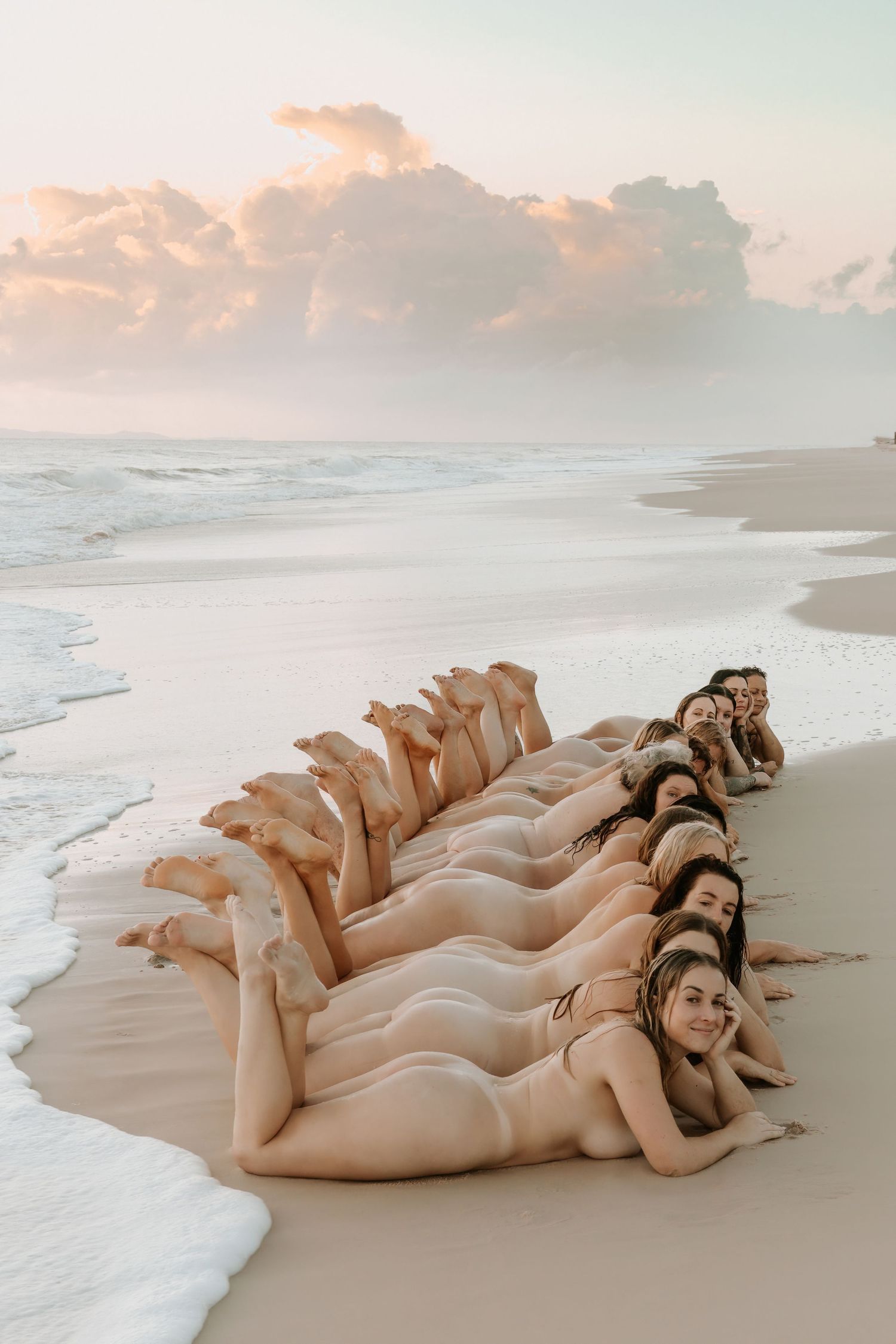 Group of women in swimsuits lying in a row on a sandy beach at sunset with gentle waves nearby.