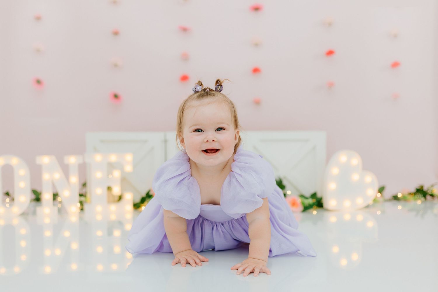 A baby in a lavender outfit crawls on the floor with twinkling string lights in the background.