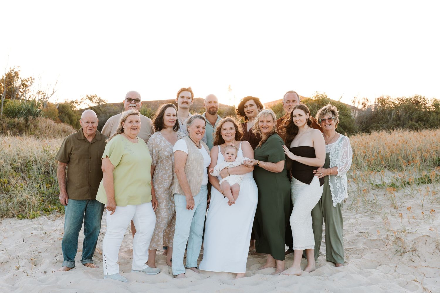 A group of people in coordinated casual outfits pose together on a sandy beach at sunset.