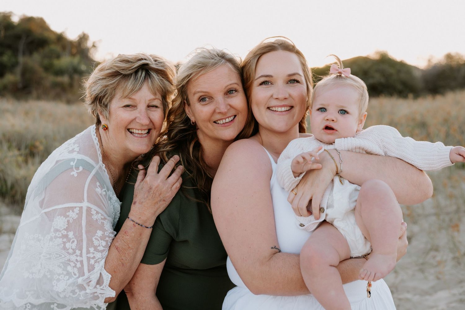 A group of people in light-colored outfits embrace and smile together during a sunset outdoor photo session.
