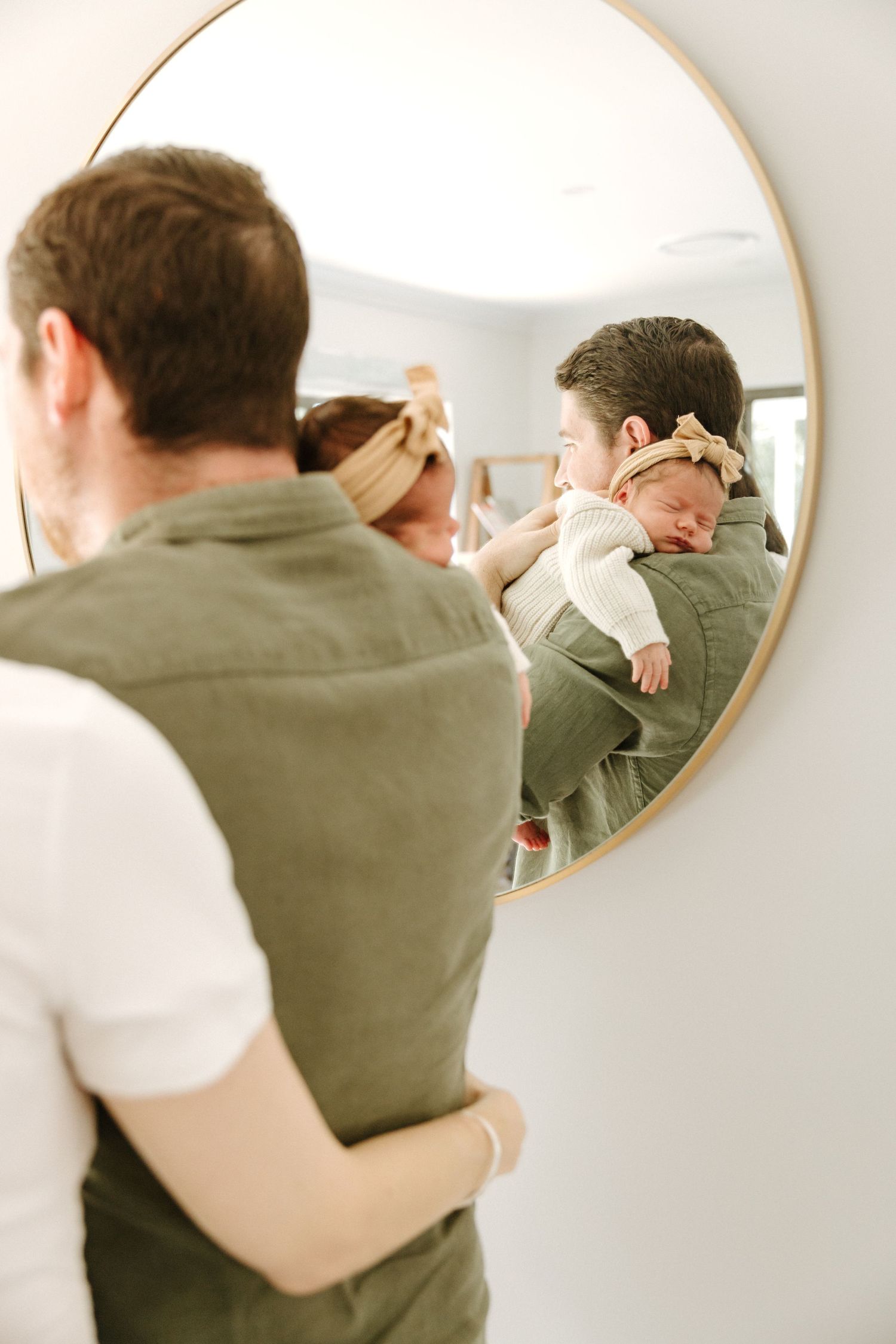 A sleeping baby is cradled against someone wearing an olive green vest, reflected in a round wall mirror.