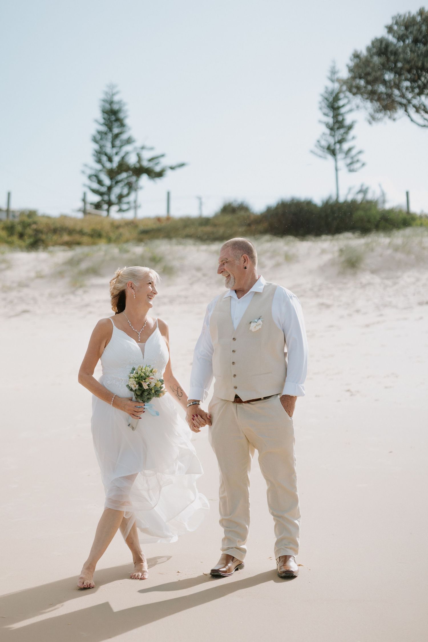 A couple walks together on a sandy beach path holding hands while dressed in white wedding attire.