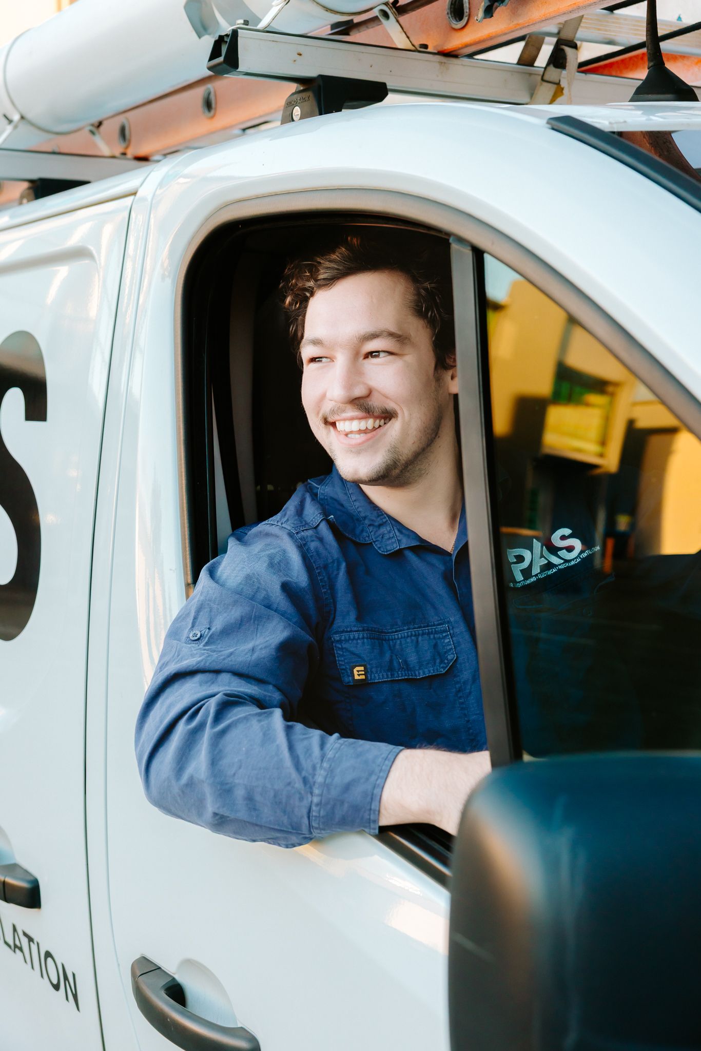 Smiling male technician in blue uniform sitting in white service van with roof rack, looking out window.
