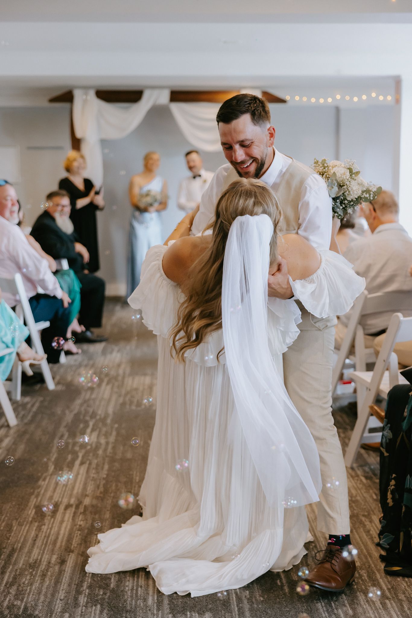 Joyful groom embracing bride in white veil as she walks down the aisle at indoor wedding ceremony.