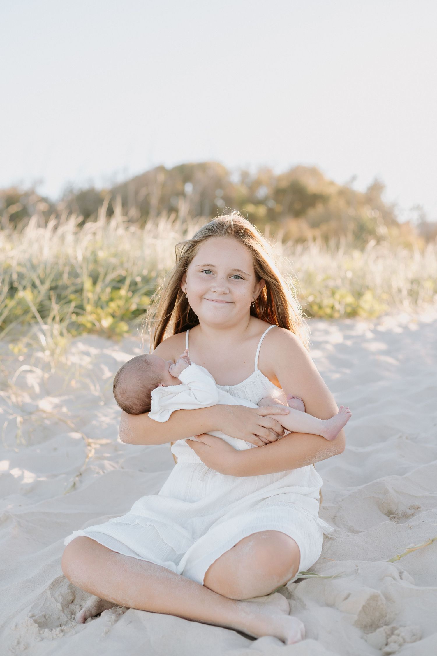 A smiling girl in a white dress sits on sandy beach dunes holding a newborn baby, with golden grass in the background.
