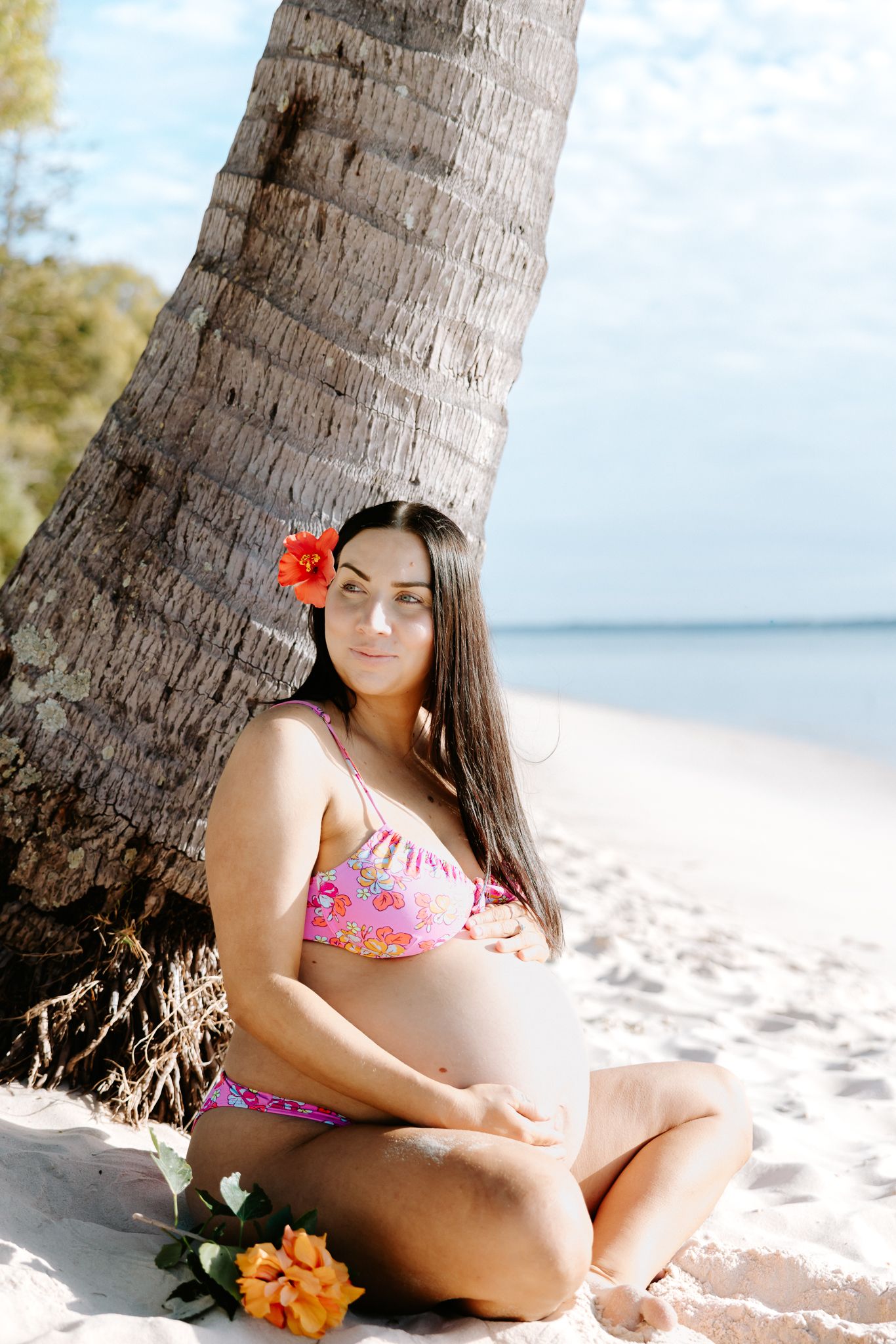 Pregnant woman in pink floral bikini sits on sandy beach leaning against palm tree with red hibiscus flower in hair.