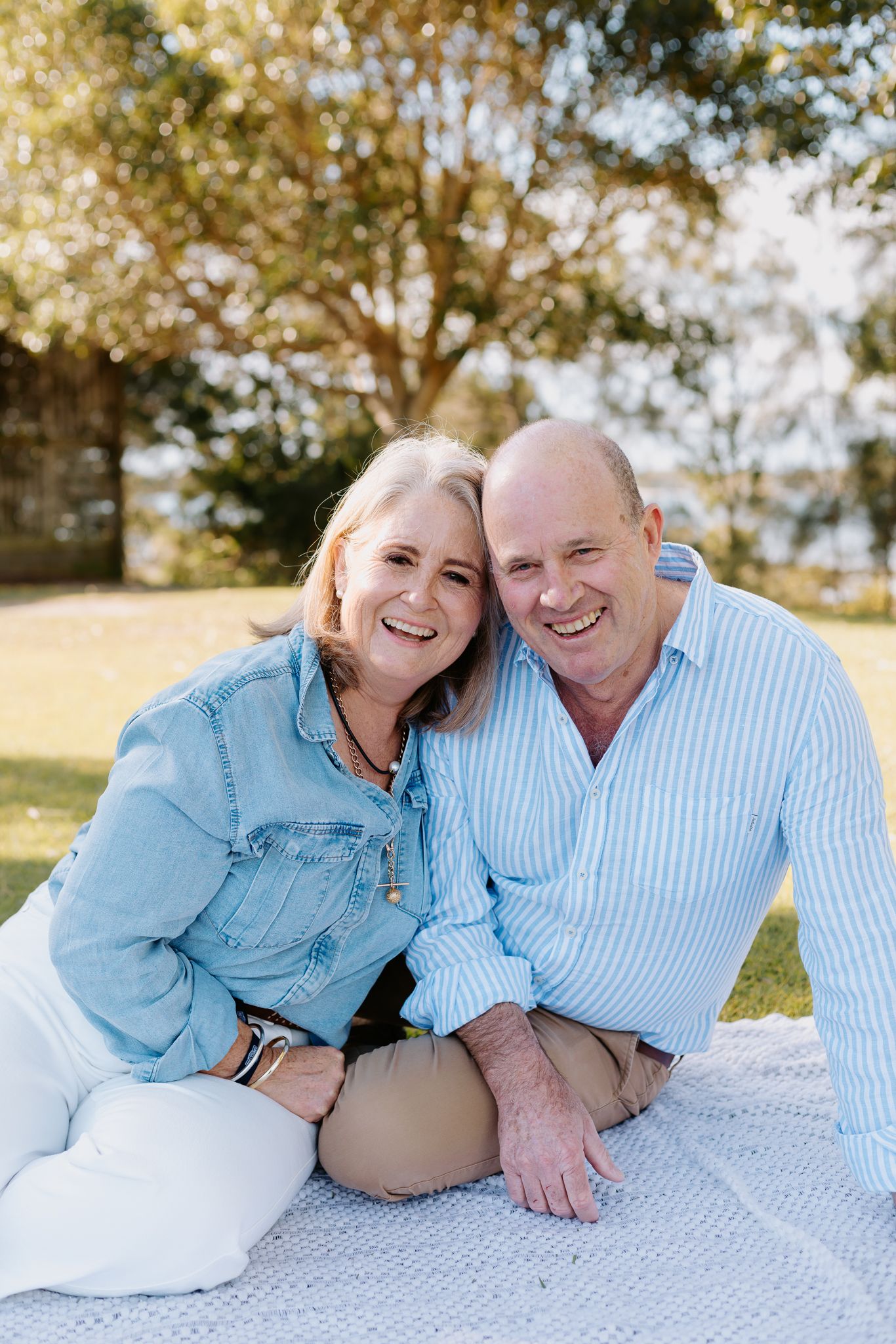 Smiling middle-aged couple sitting on a blanket outdoors in a sunny park, wearing light blue casual outfits.