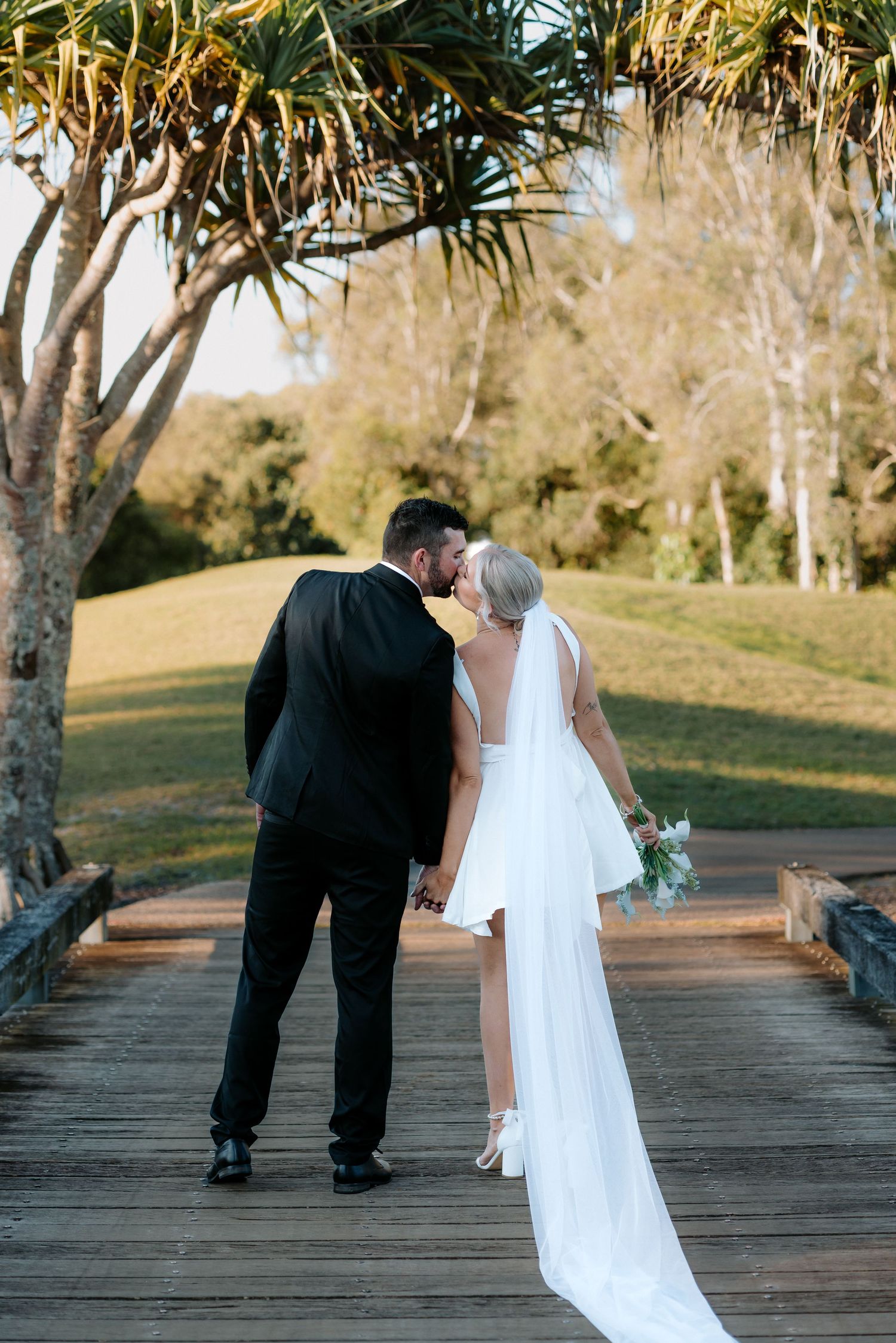 Bride and groom sharing a kiss on a wooden bridge surrounded by lush greenery on their wedding day.