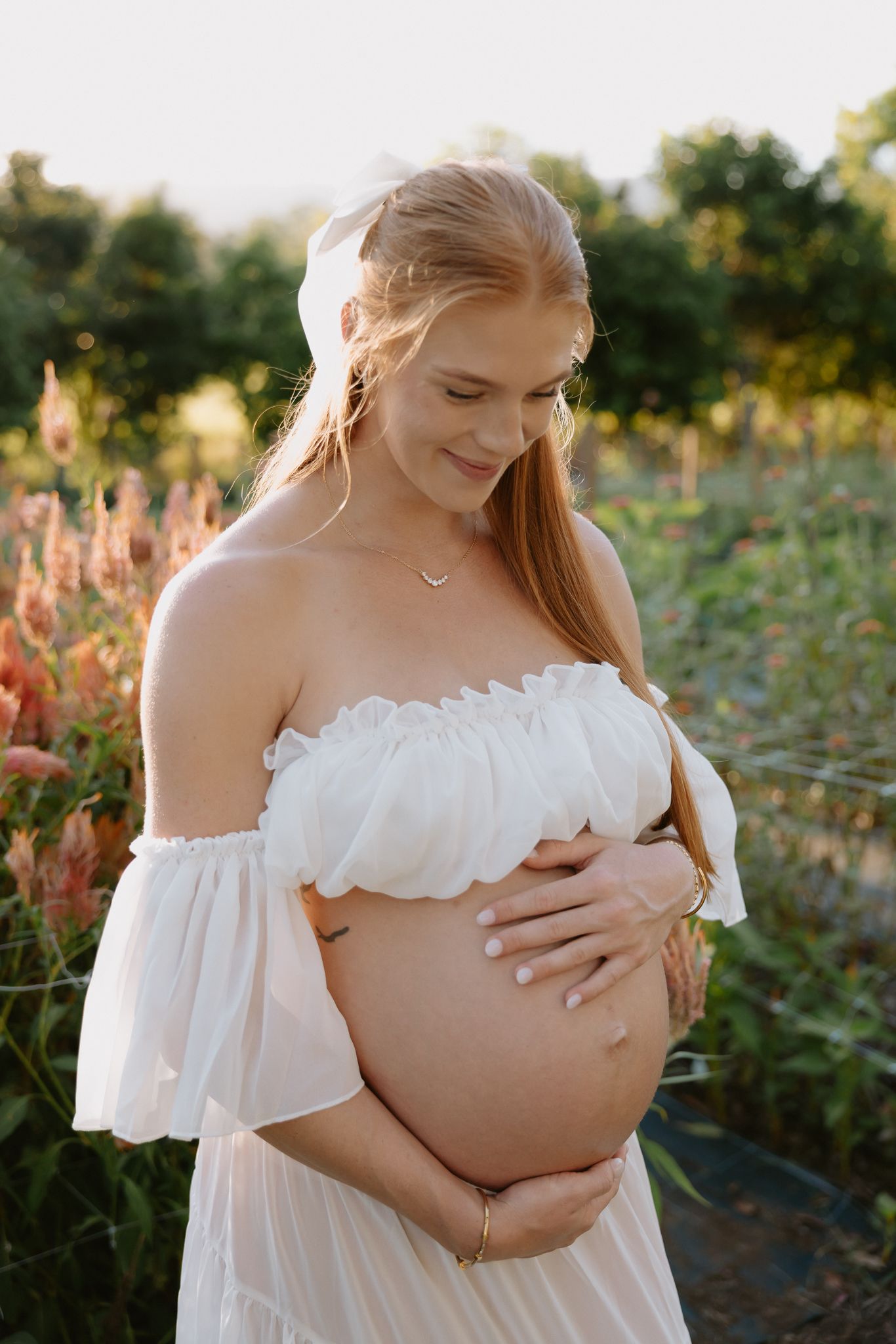 Pregnant woman in white off-shoulder dress holding baby bump outdoors in golden hour light among pink flowers.