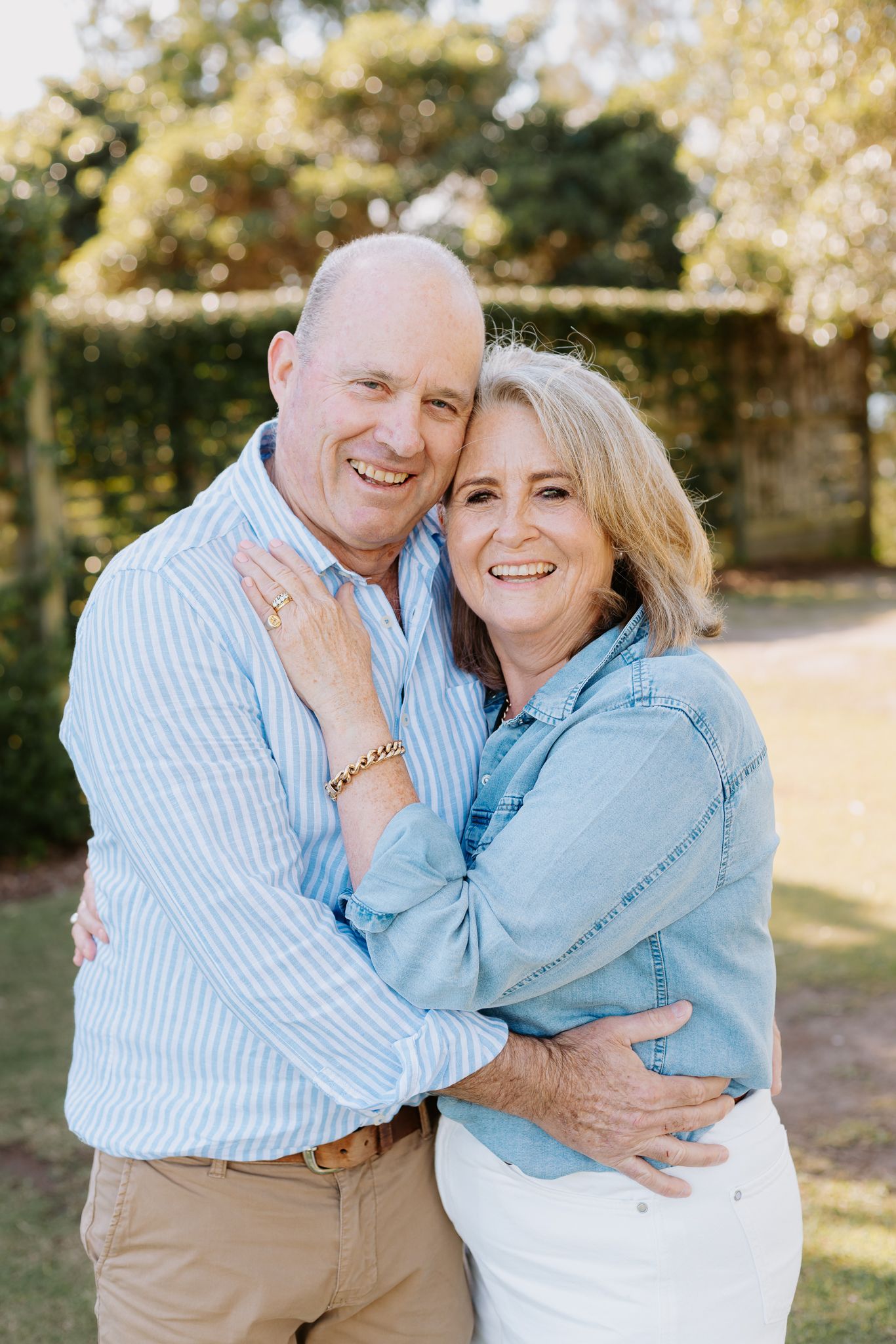 Happy middle-aged couple embracing outdoors, smiling warmly in casual blue shirts against a green garden backdrop.