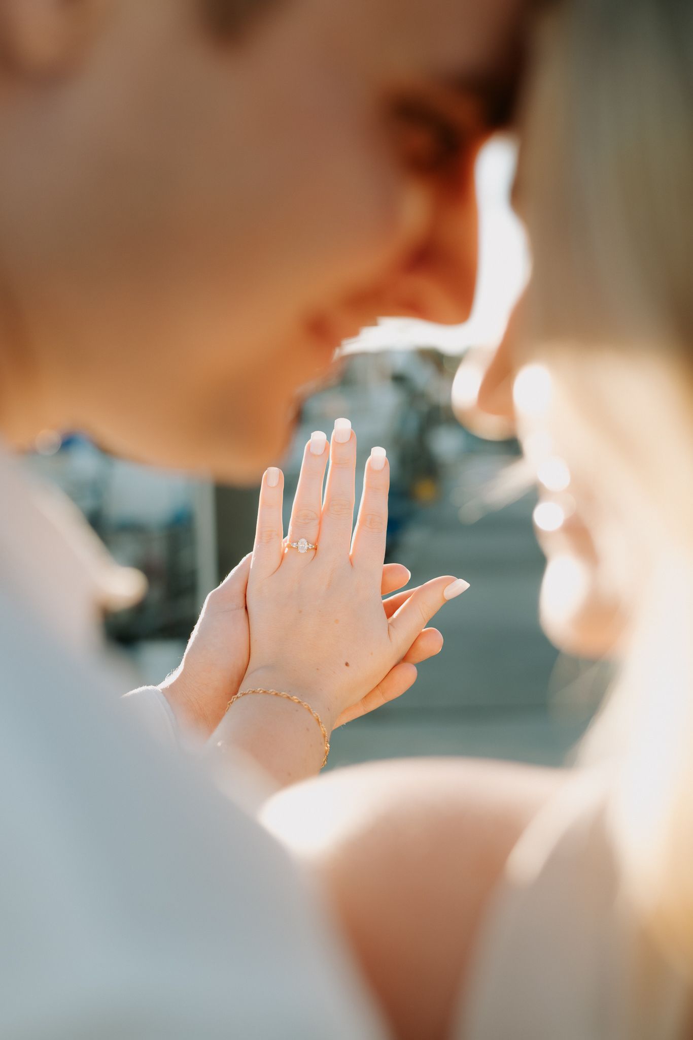 Couple sharing a kiss while showing off an engagement ring with a diamond solitaire and delicate bracelet in warm sunlight.
