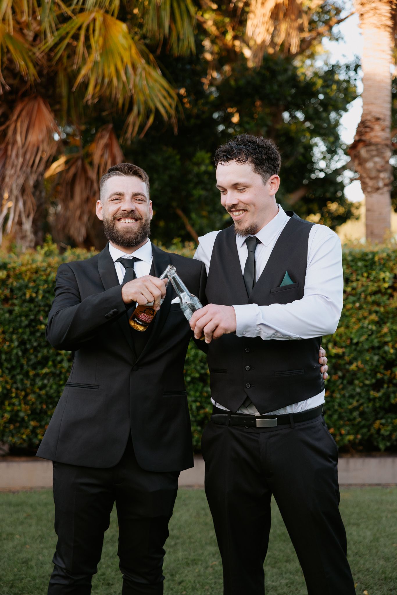 Two well-dressed grooms in black suits clink beer bottles outdoors near palm trees at their wedding celebration.