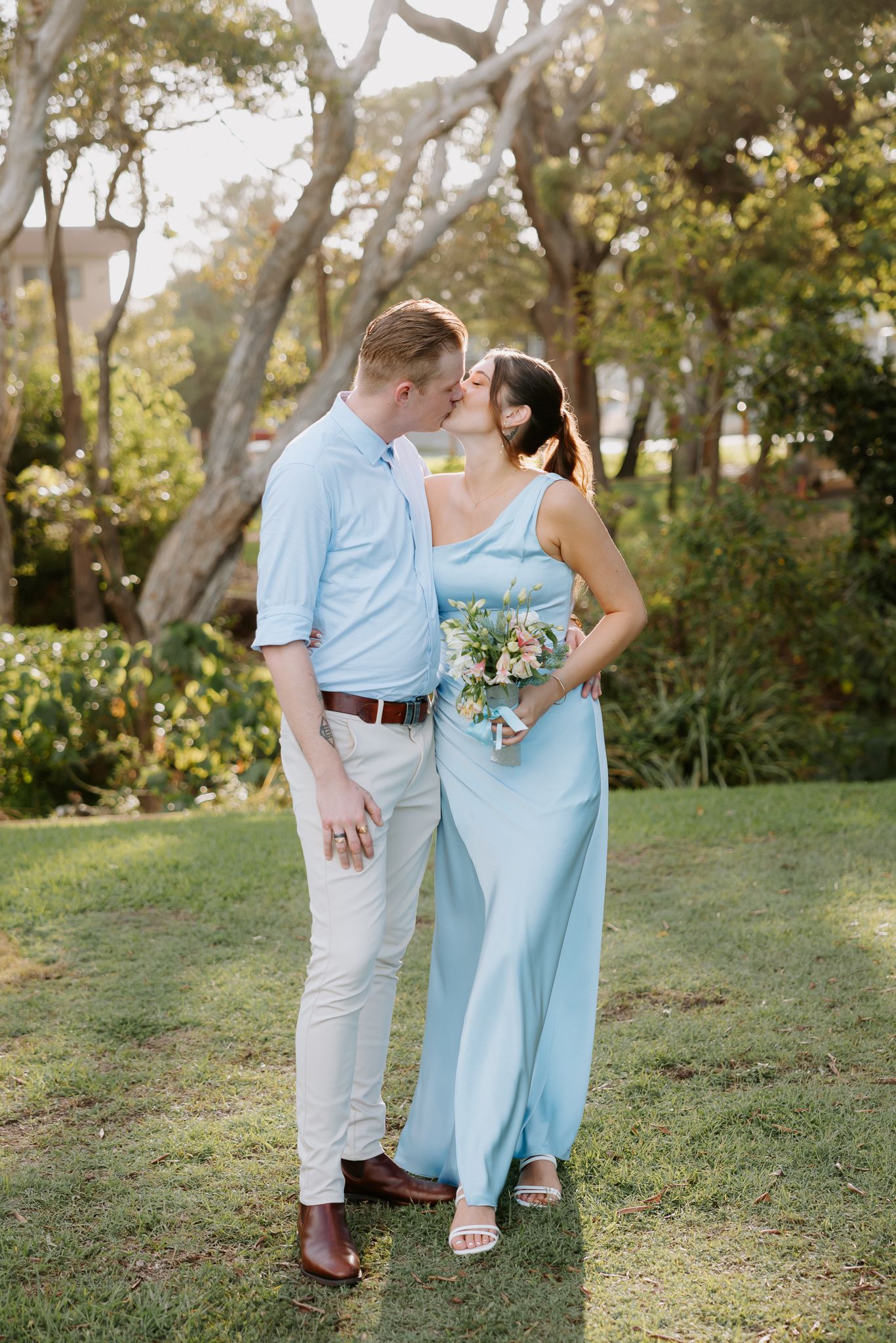 Couple kissing in garden, woman in light blue maxi dress holding bouquet, man in blue shirt and white pants.