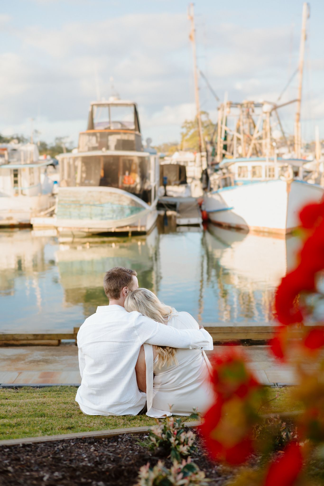Couple sitting together at a marina dock, surrounded by boats and red flowers, enjoying a peaceful waterfront moment.