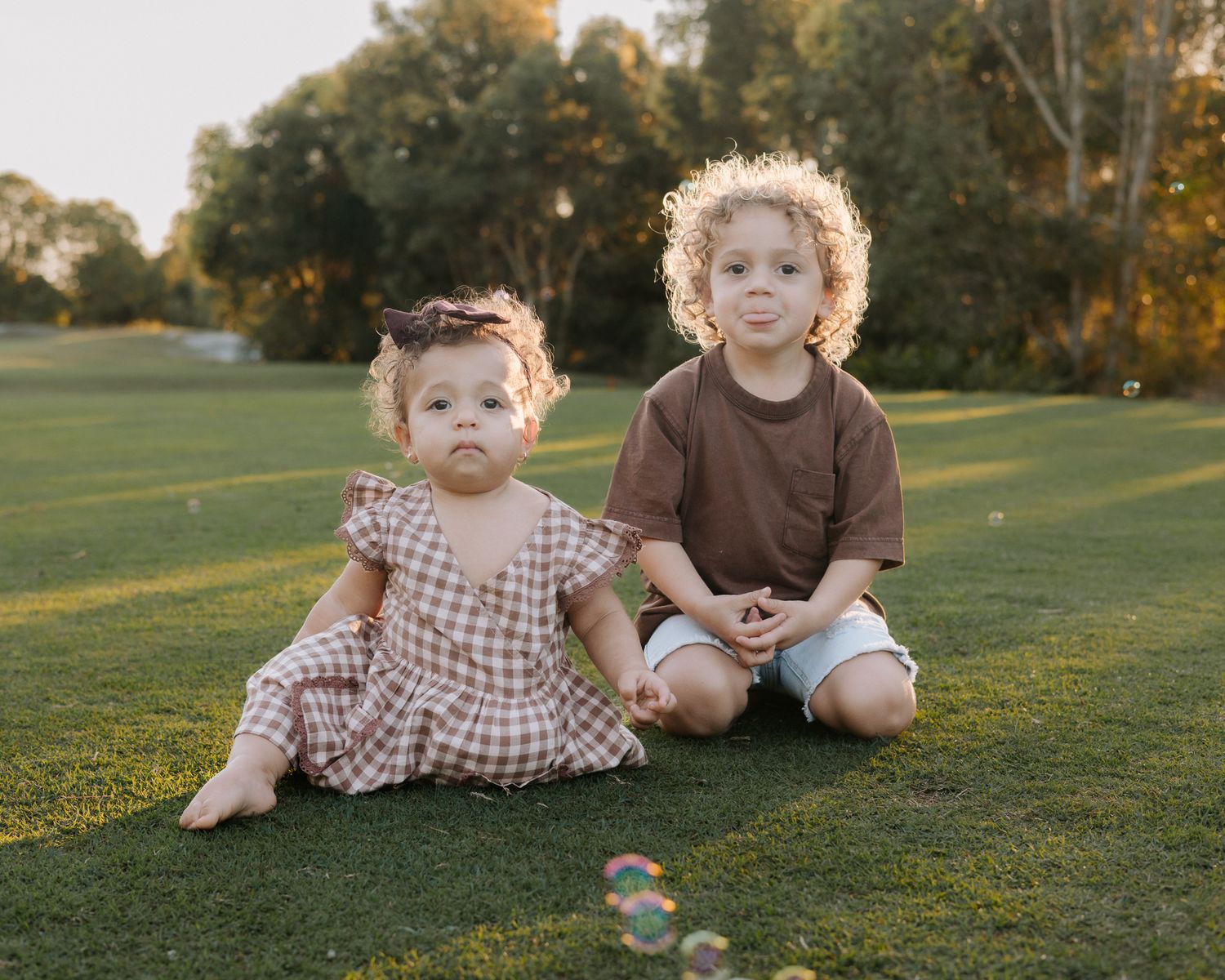 Two young children sitting on green grass in a sunny park, girl in plaid dress and boy in brown shirt with bubbles.