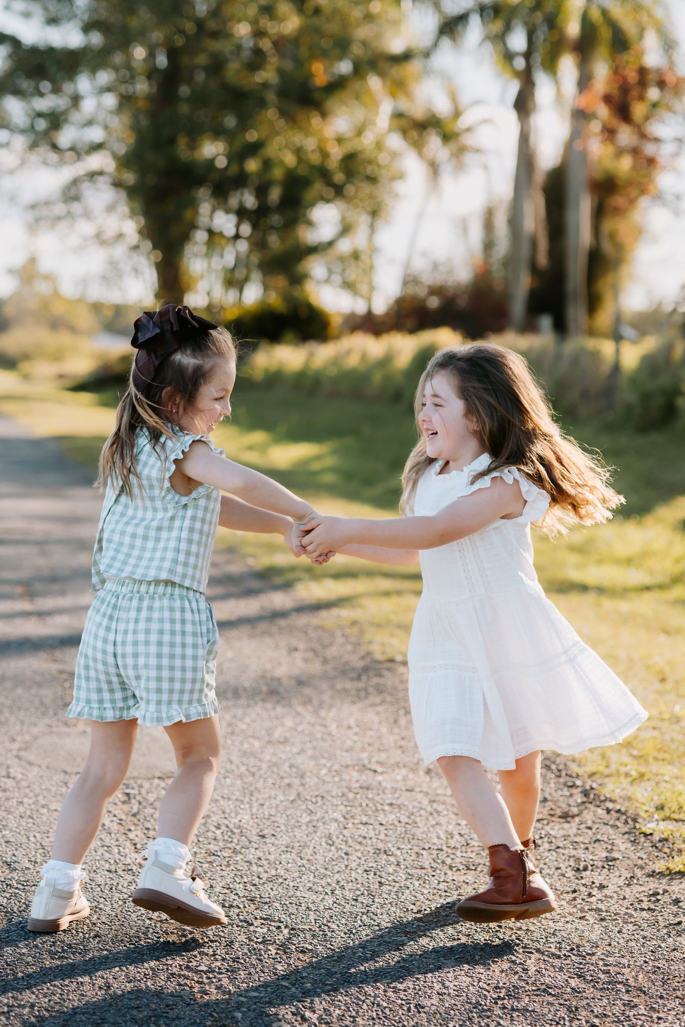 Two young girls in dresses hold hands and spin together on a sunny outdoor path surrounded by green trees.