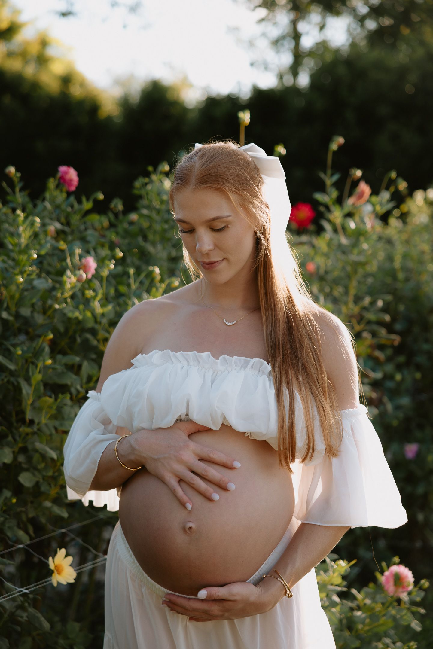 Pregnant woman in white off-shoulder dress holding baby bump, surrounded by blooming roses in golden hour light.