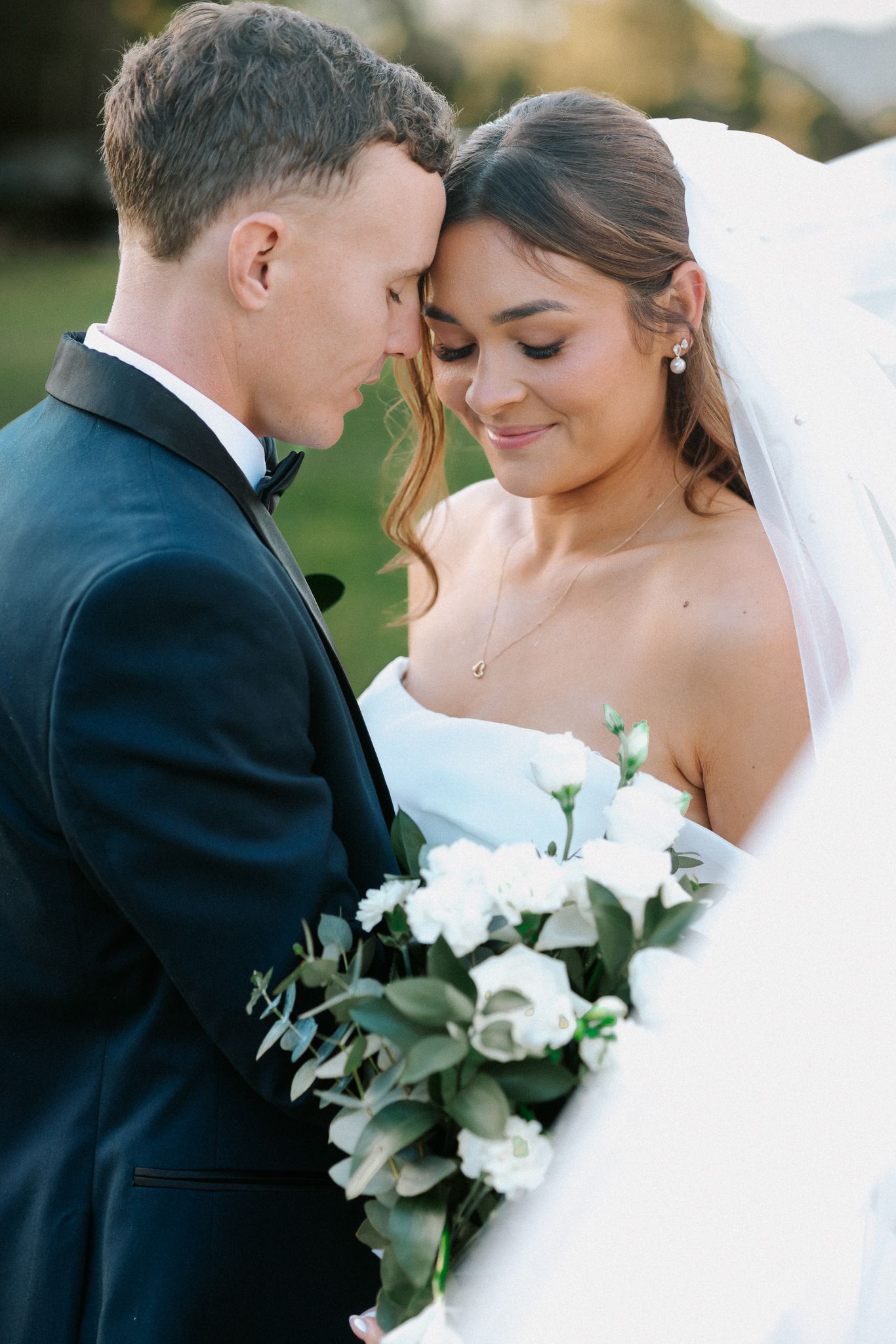 Close-up of smiling bride in white gown with veil holding white roses, touching foreheads with groom in black tux.