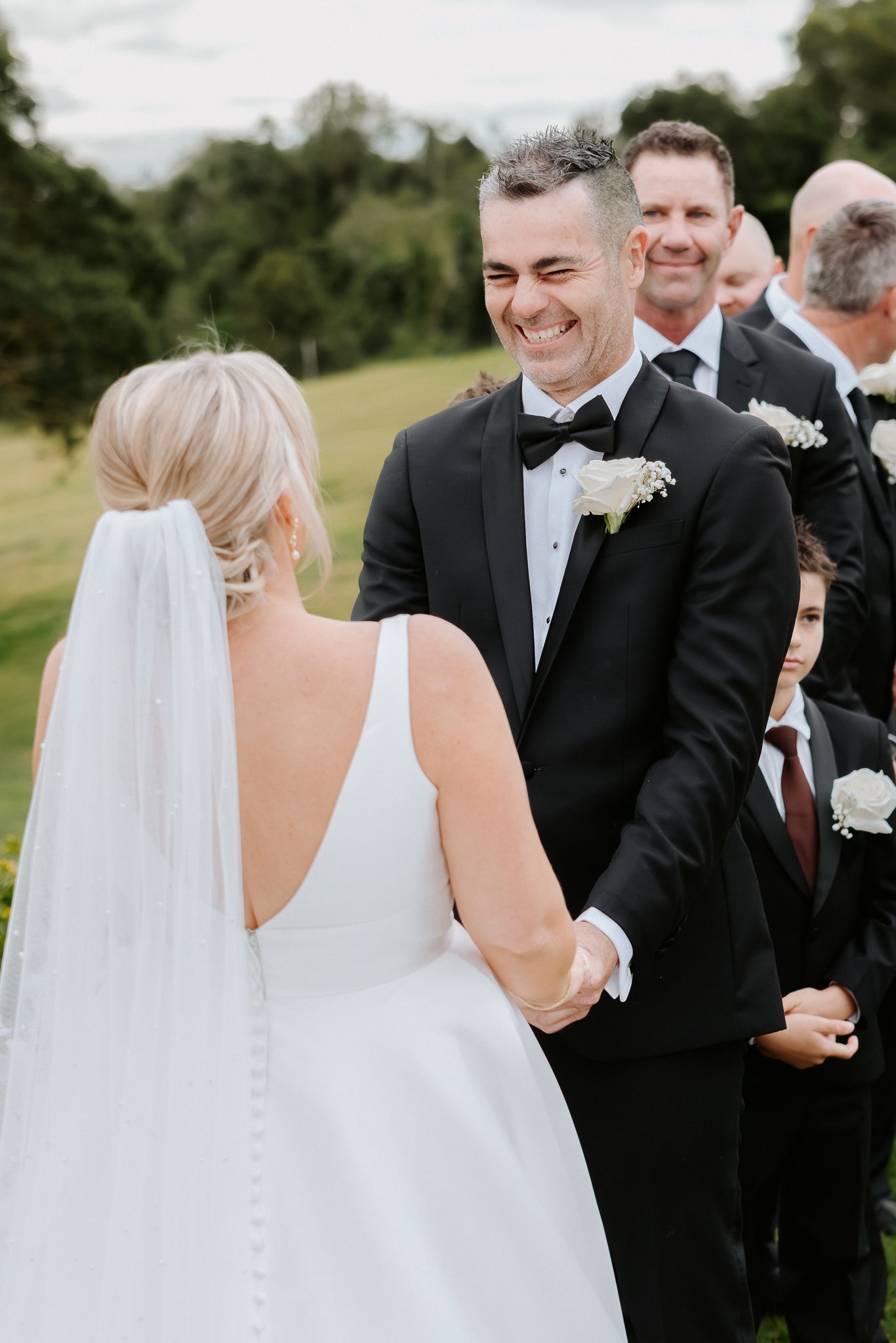 Groom in black tuxedo smiling joyfully at bride in white wedding dress during outdoor ceremony.