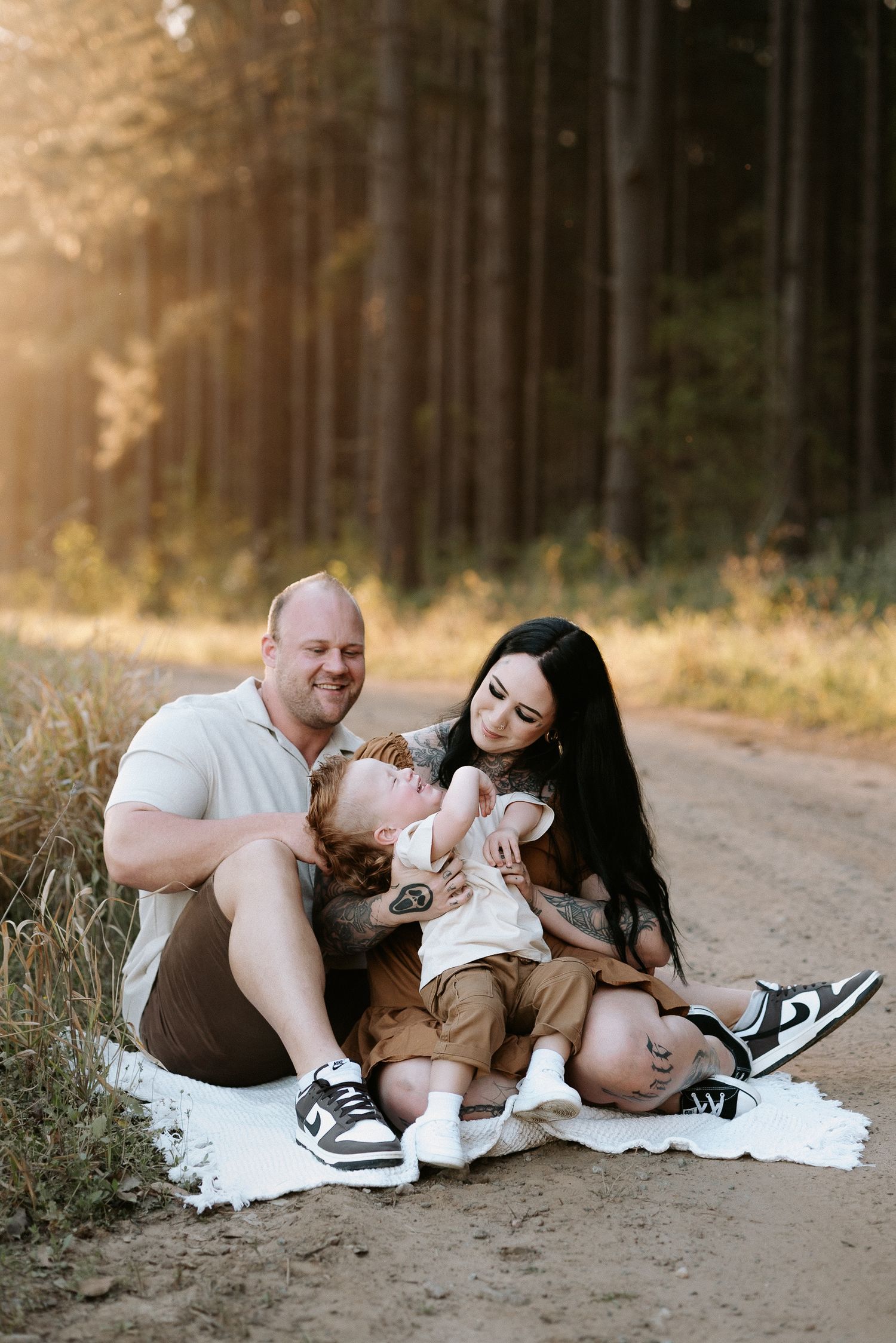 Family sitting on blanket on forest dirt road at golden hour, parents smiling at laughing toddler.