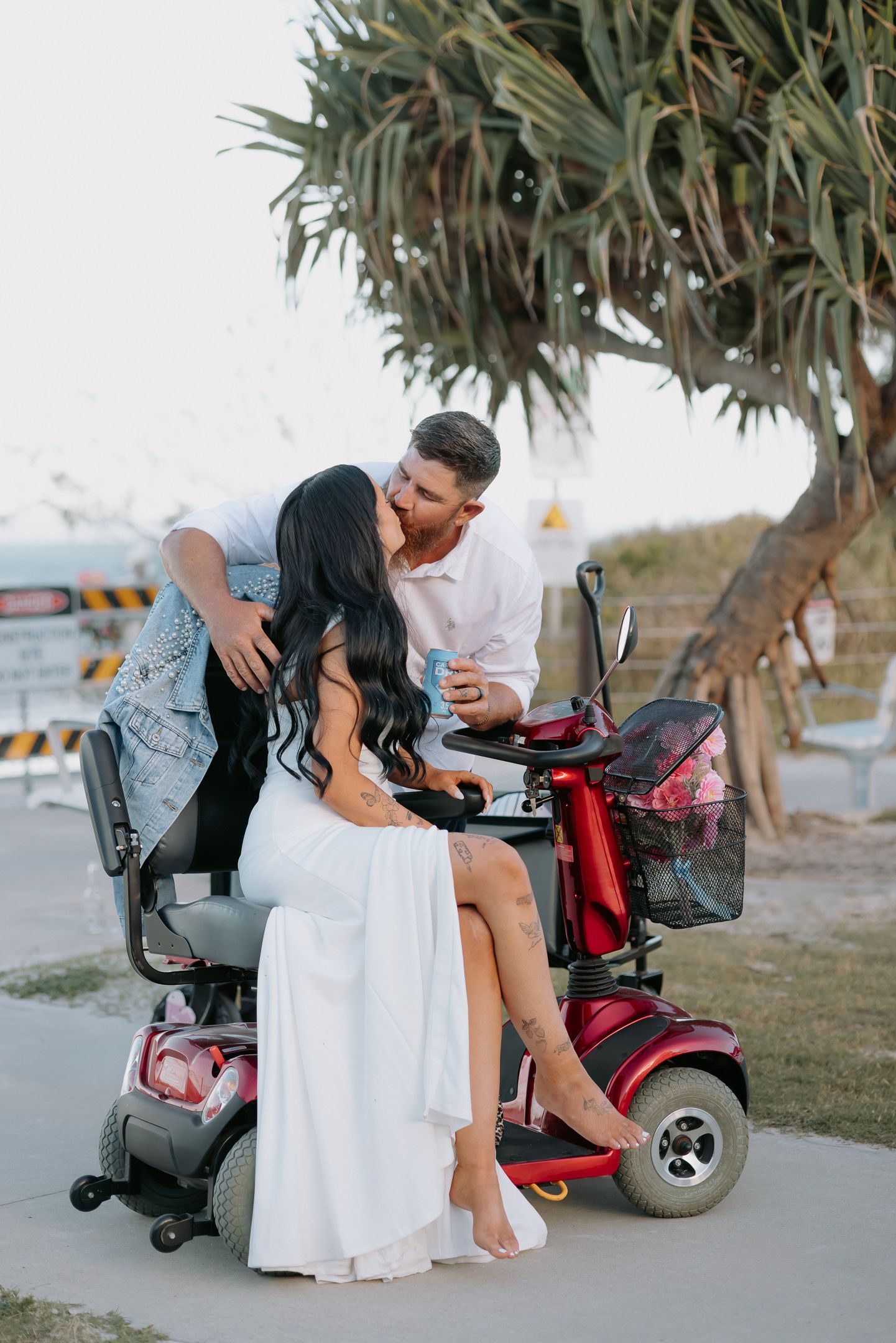 A romantic couple shares an intimate moment on a mobility scooter by the beach under a palm tree at sunset.