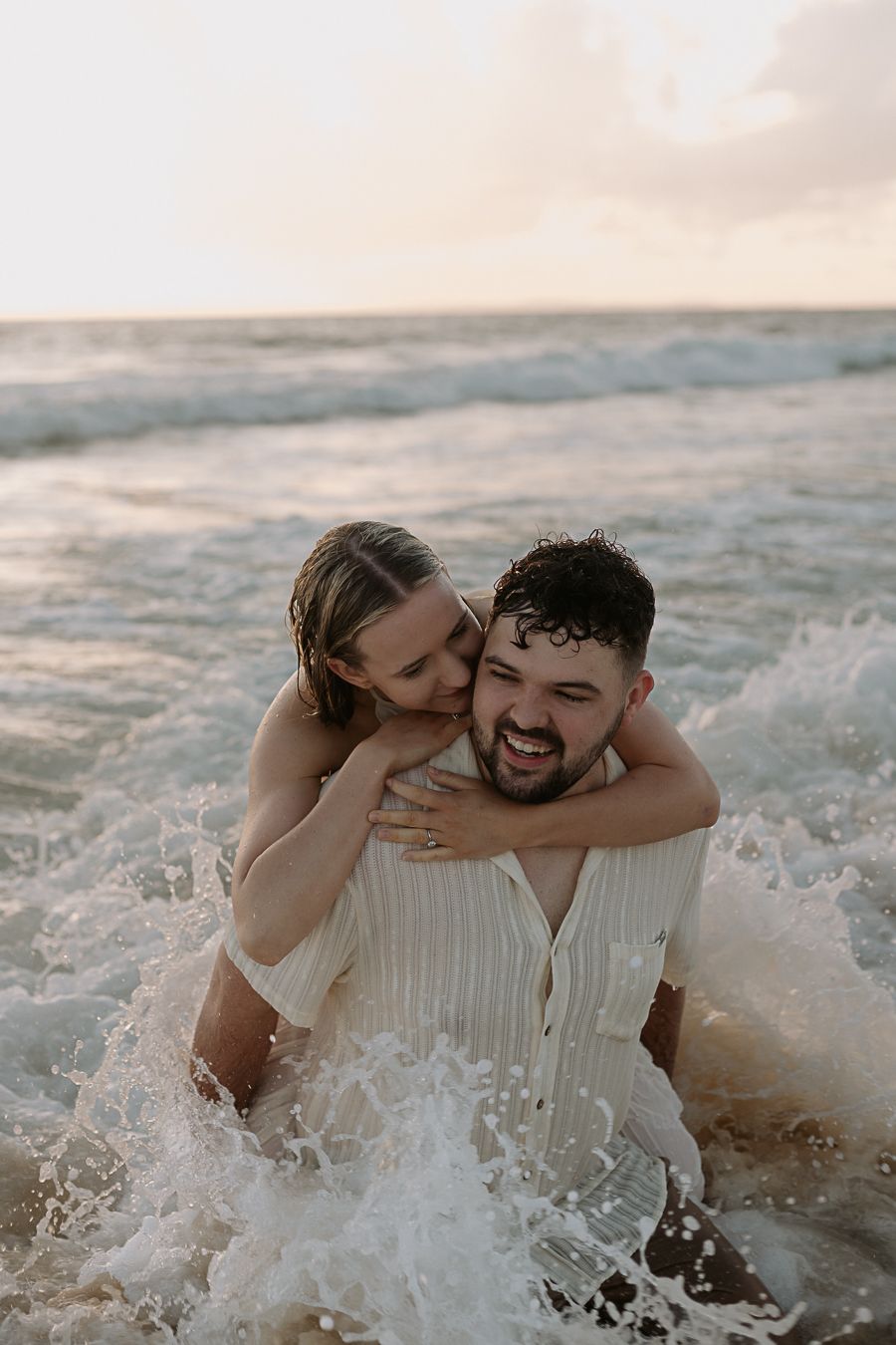 Couple laughing and embracing in ocean waves at sunset, wearing white outfits during a romantic beach photo session.