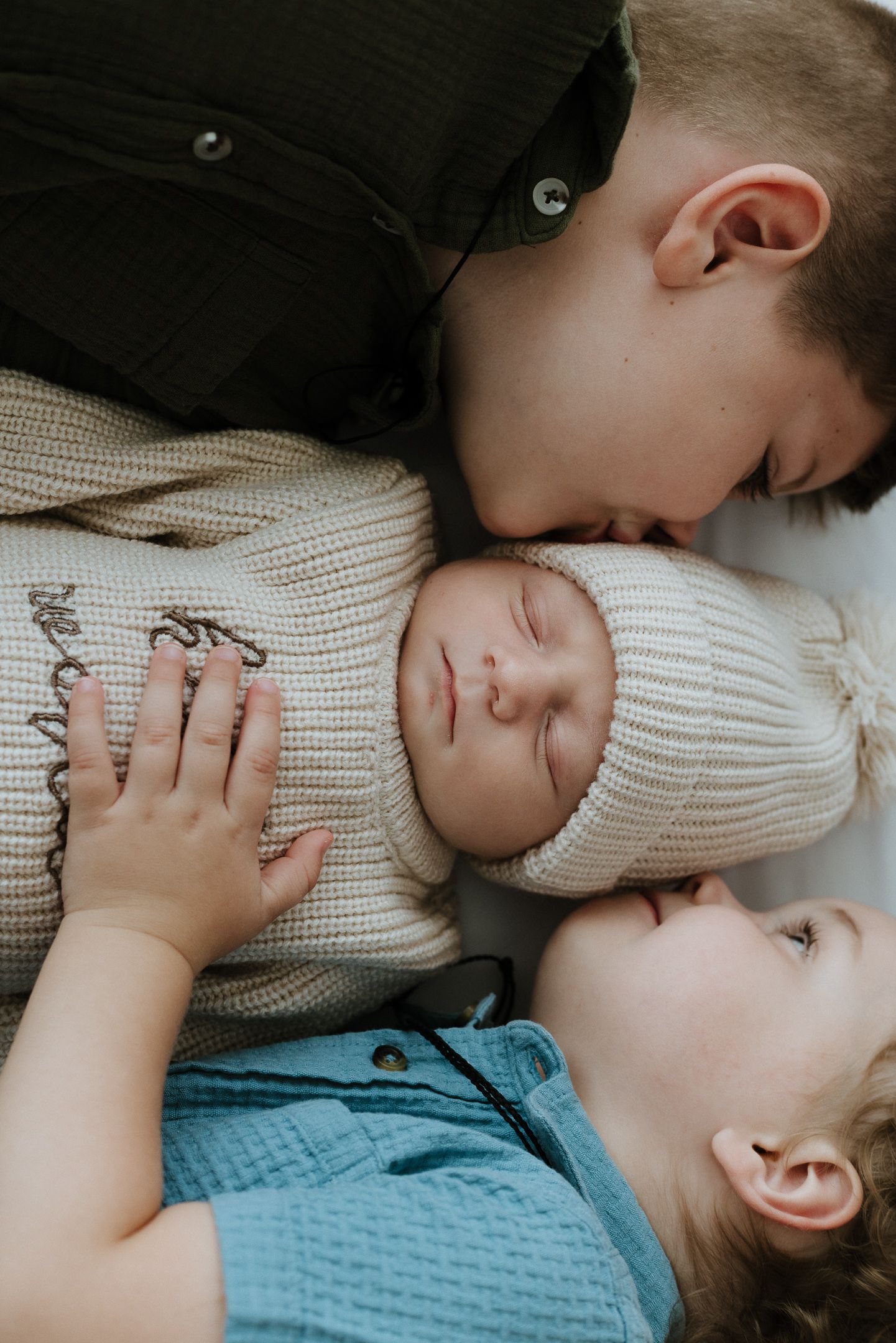Three siblings lying close together in cozy knit sweaters sharing a tender family moment.